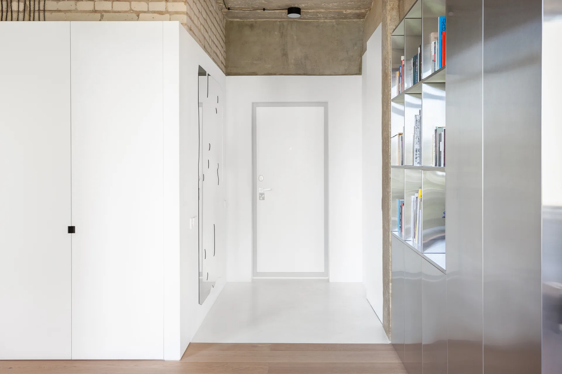 A minimalist entryway hallway features white built-in closets, a reflective metallic shelving unit, and an exposed raw concrete ceiling.