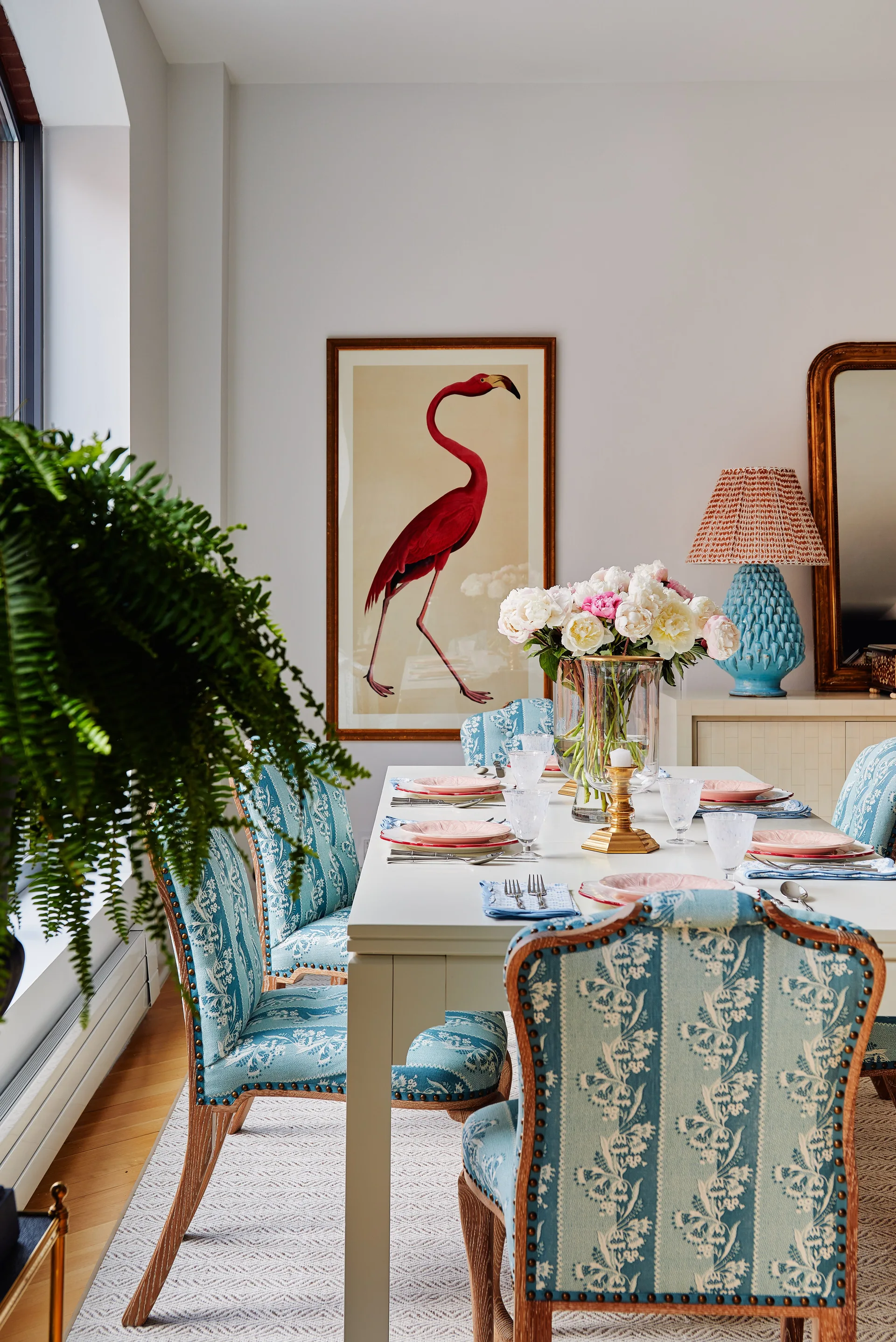 A white rectangular dining table set for a meal is paired with blue and white botanical patterned upholstered chairs in a bright room.