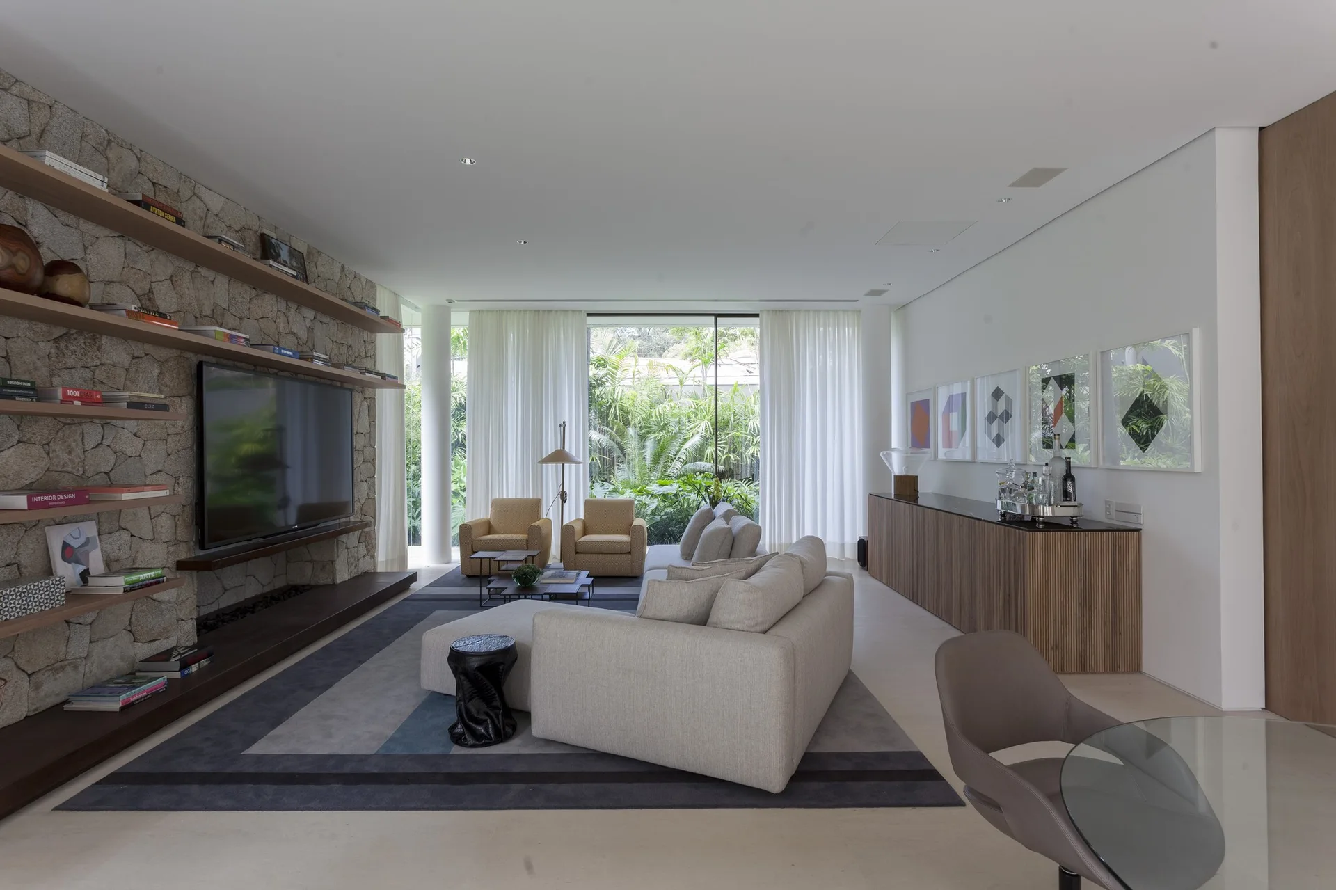 A modern living room features a stacked stone accent wall with floating wooden shelves, a light gray sectional sofa, and floor-to-ceiling windows.