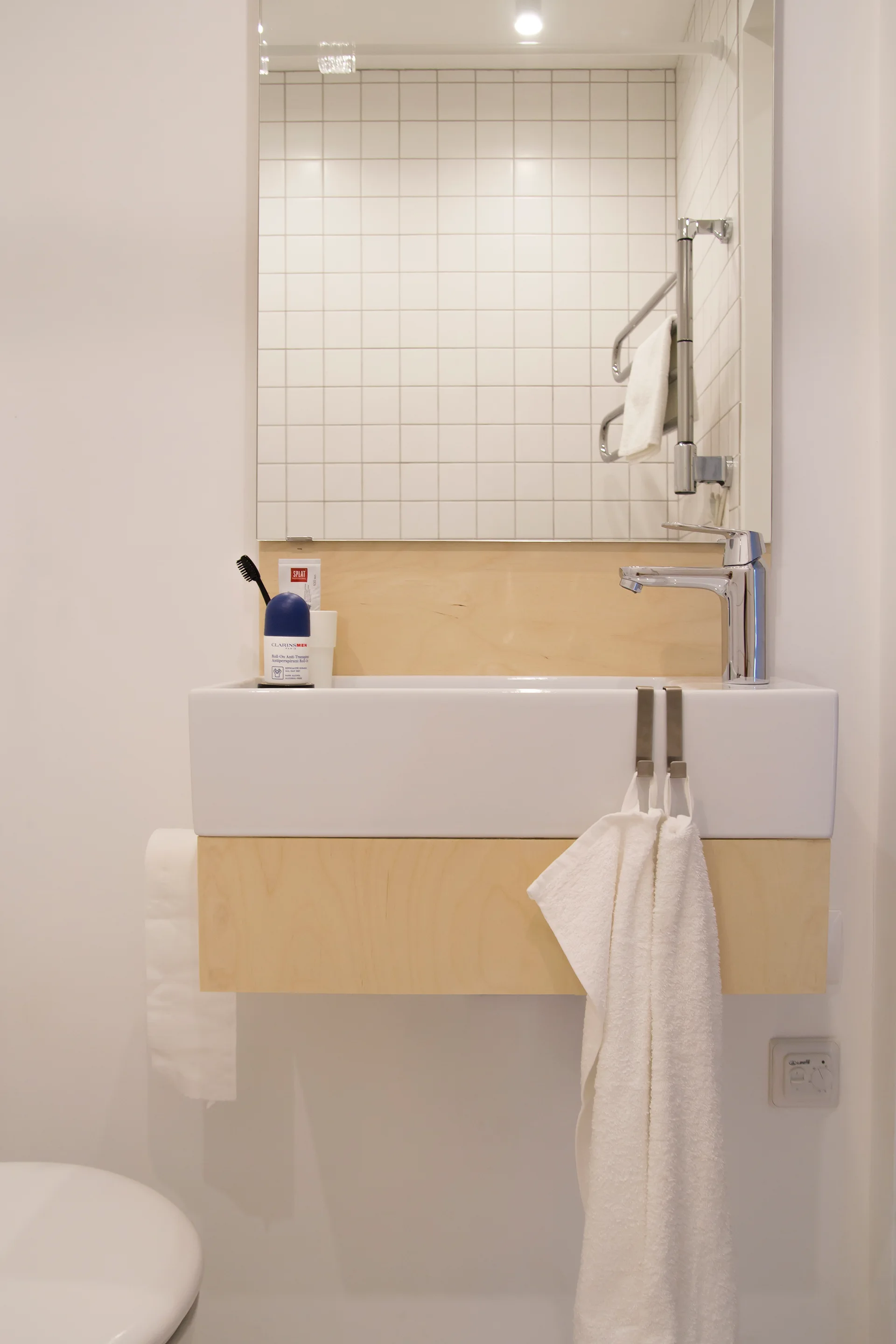 A wall-mounted white ceramic sink features a light wood under-panel and is set against a white grid-tiled wall beneath a rectangular mirror.