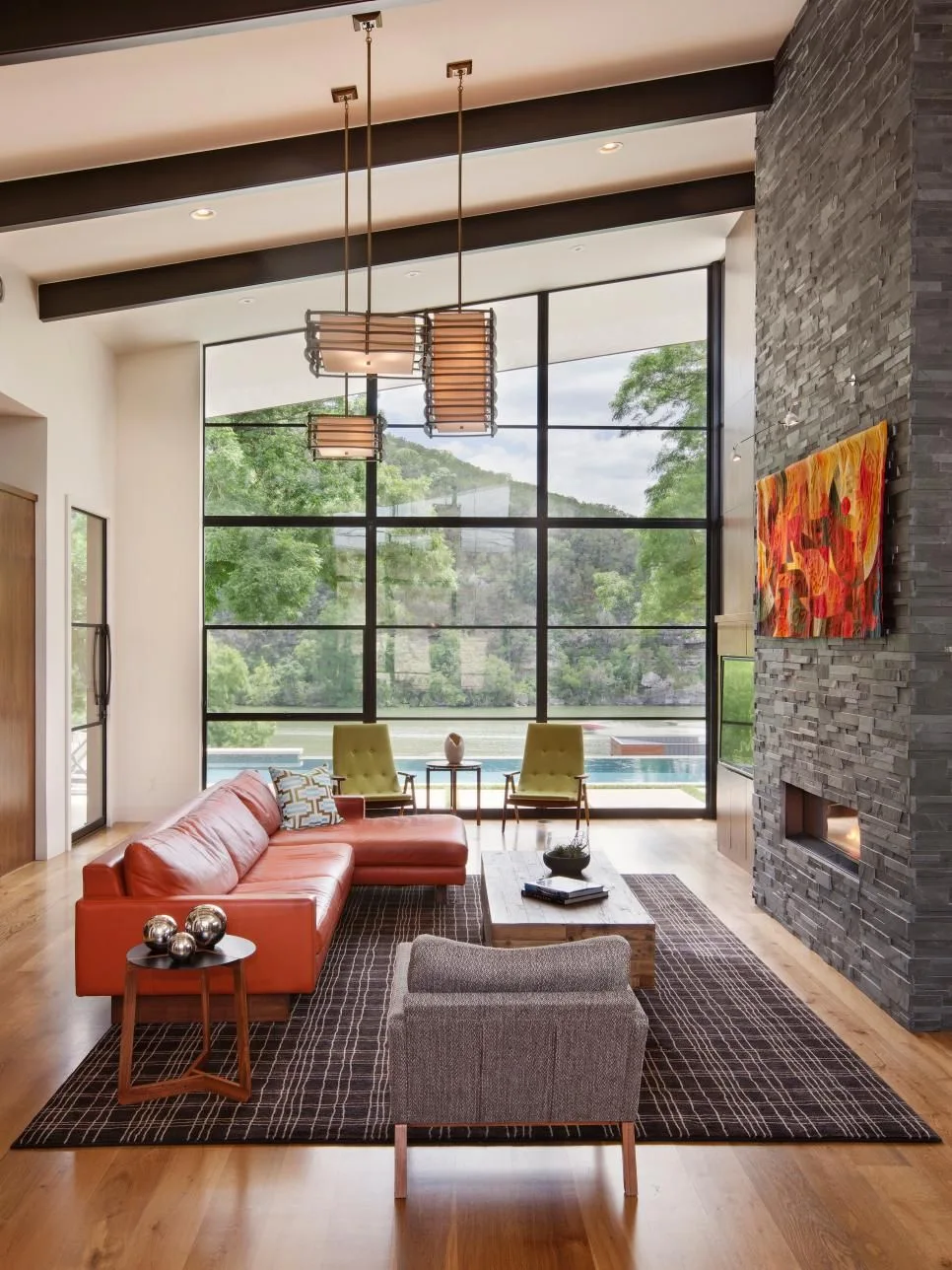 A modern living room with an orange leather sectional, a textured grey armchair, a tall stone fireplace, and a large grid window showing a river.