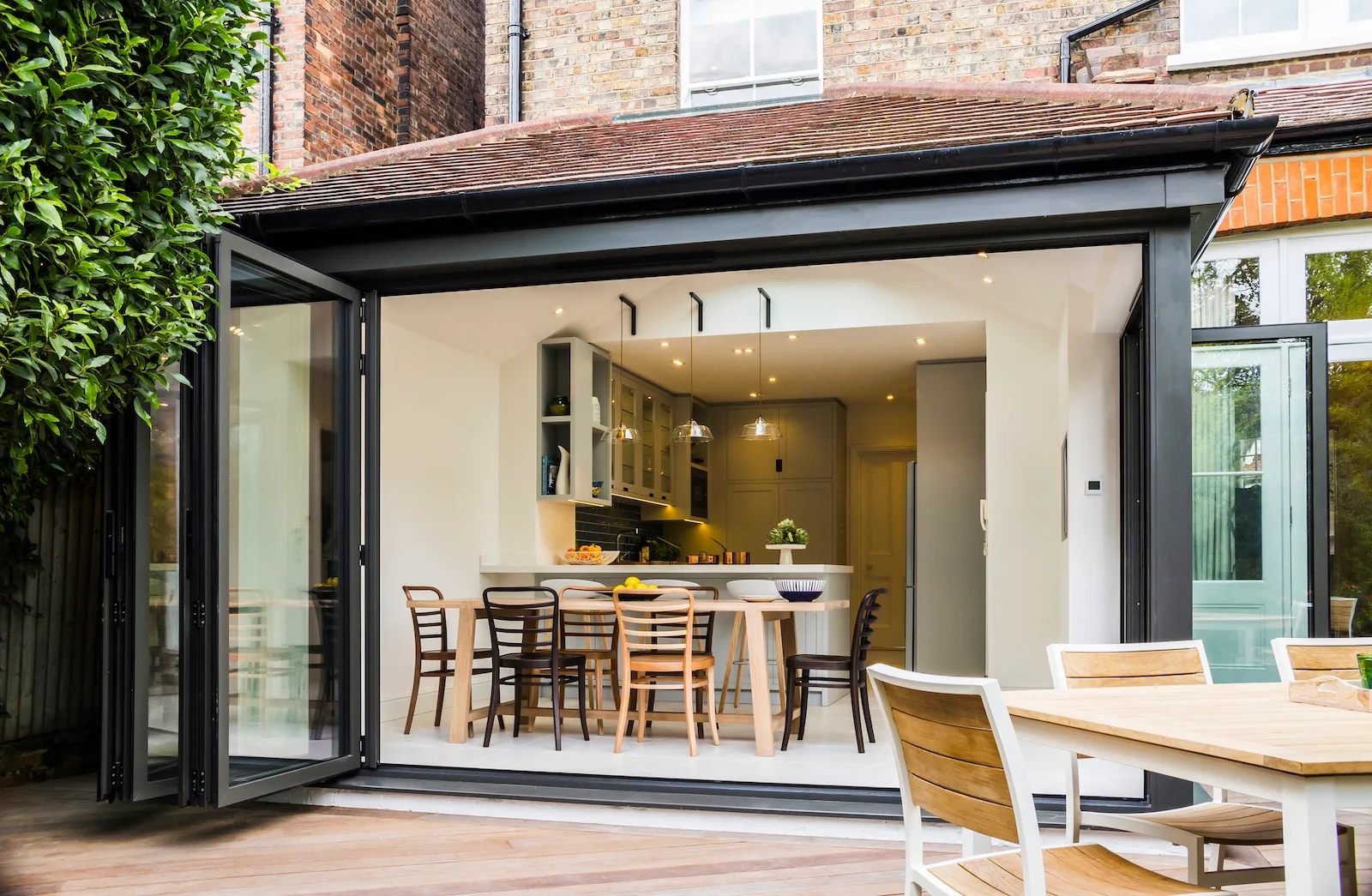 A modern kitchen and dining area with light wood table and chairs opens onto a wooden deck through large black bi-fold glass doors.