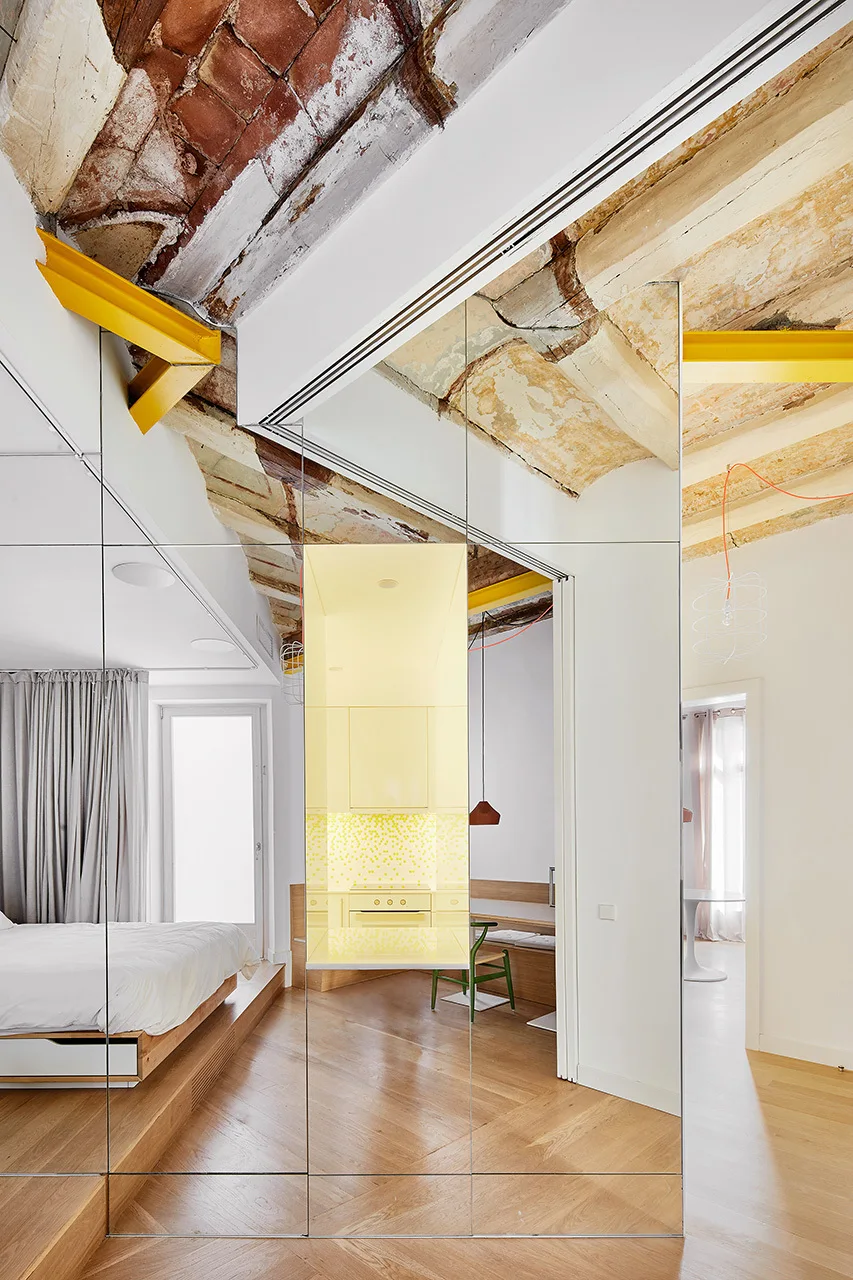 A wooden platform bed sits beside a tall mirrored wall reflecting a yellow kitchen under an exposed brick and mortar ceiling with yellow beams.