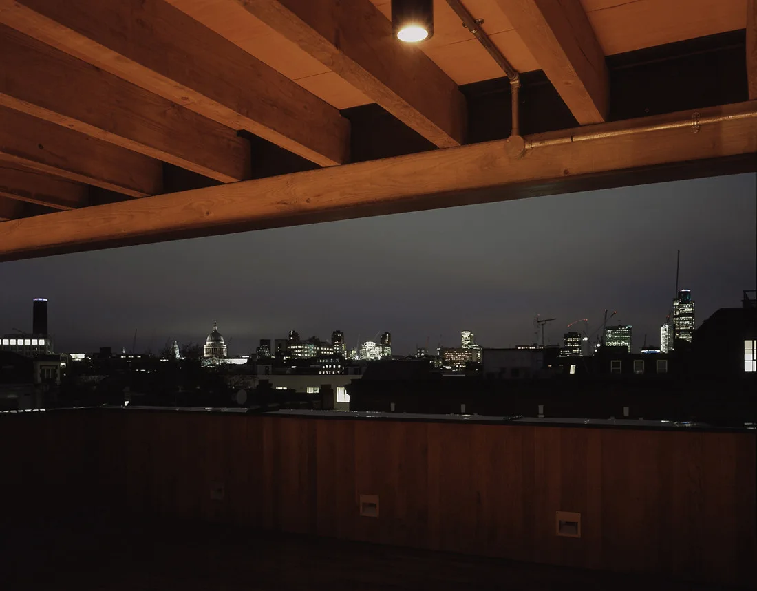 An outdoor roof terrace at night featuring exposed timber ceiling beams, a wood-paneled railing, and a distant city skyline view under a dark sky.