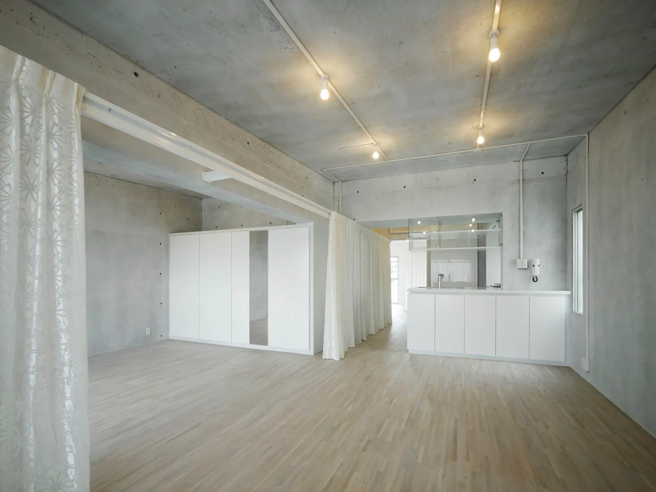 A minimalist apartment interior features raw concrete walls and ceiling, light wood floors, sleek white cabinets, and sheer white curtains.