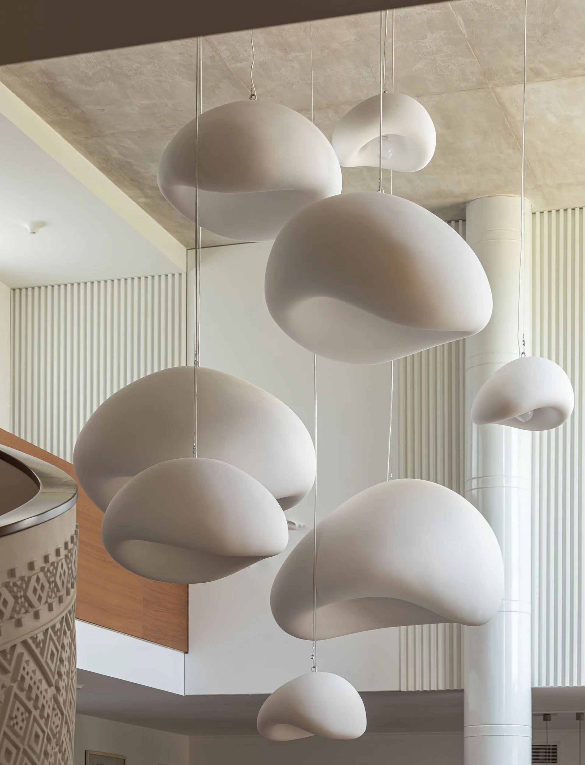 A cluster of white organic-shaped pendant lights hangs at varying heights in a stairwell with textured vertical wall paneling and a curved balustrade.