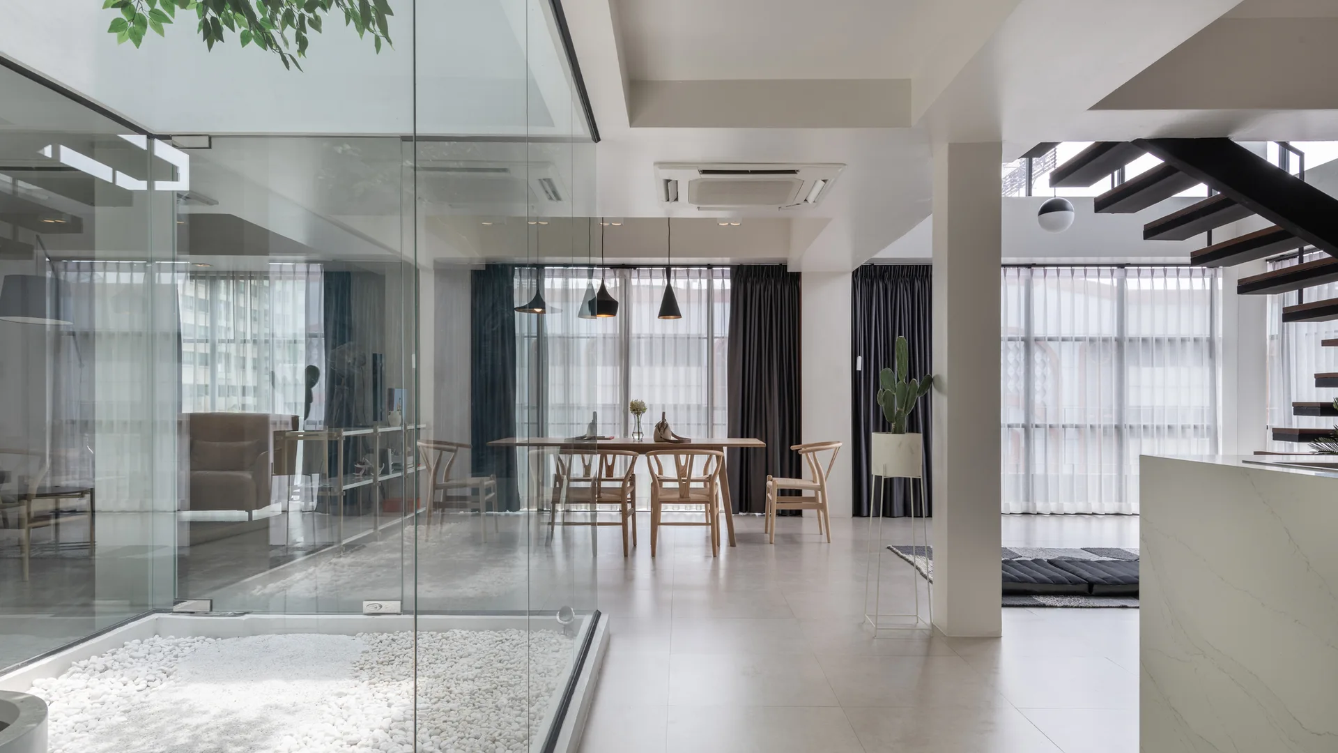 A minimalist dining room features a wooden table, wishbone chairs, and a glass-enclosed indoor courtyard with white pebbles.
