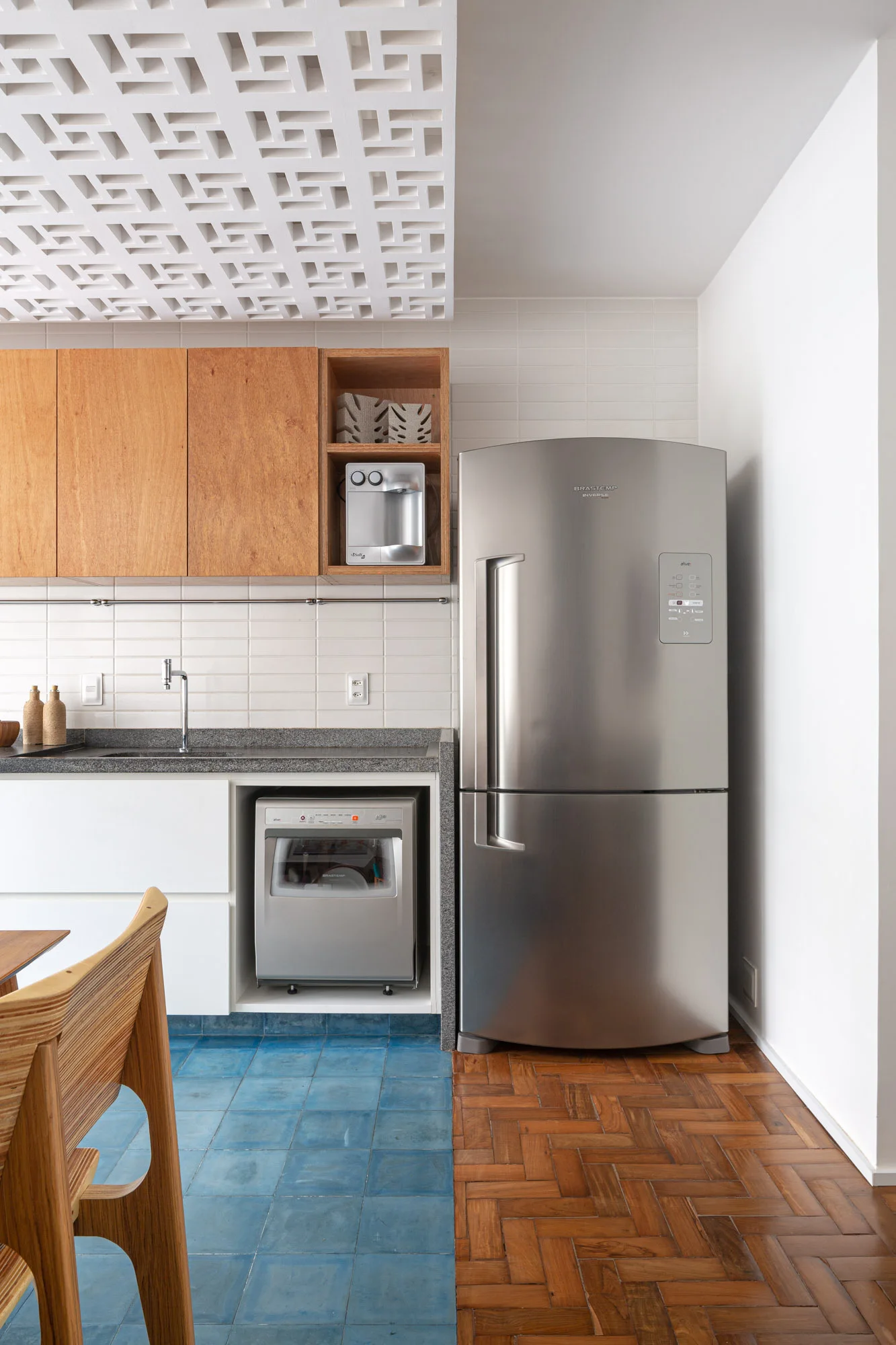 A modern kitchen shows a stainless steel refrigerator, white and wood cabinetry, and a floor transitioning from blue tiles to herringbone wood.