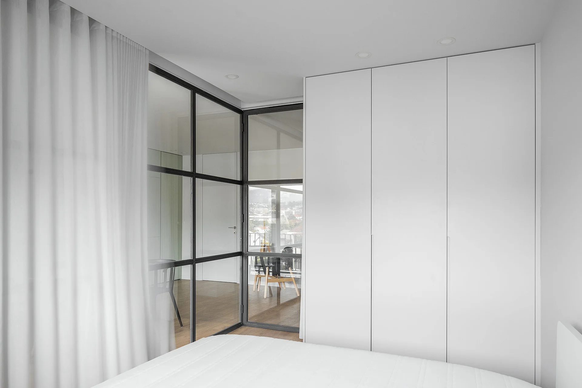 A minimalist white bedroom features a built-in wardrobe and a black-framed glass partition wall next to a sheer curtain.