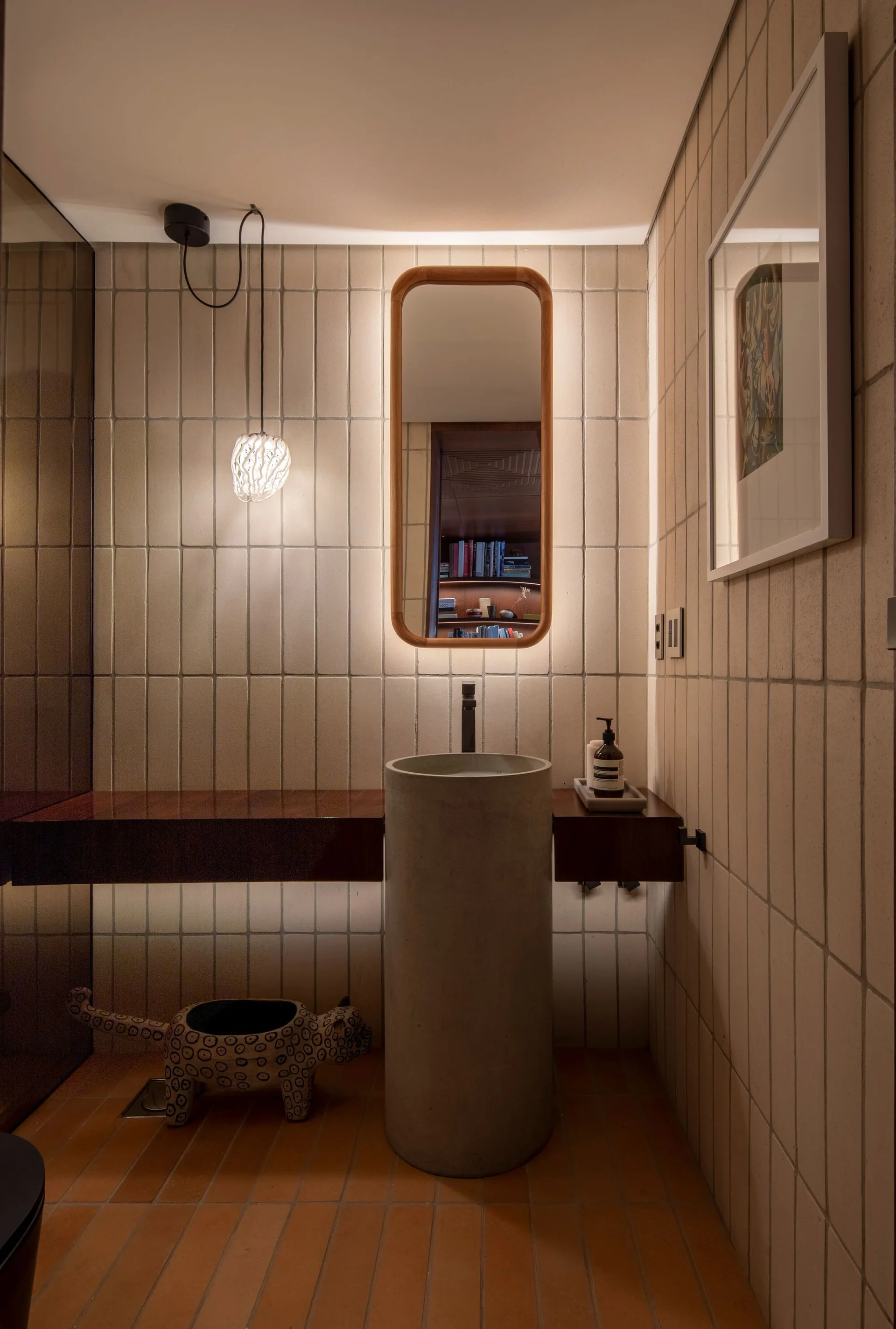 A modern powder room features a cylindrical stone pedestal sink, a wood-framed backlit mirror, vertically stacked cream tiles, and terracotta floor tiles.