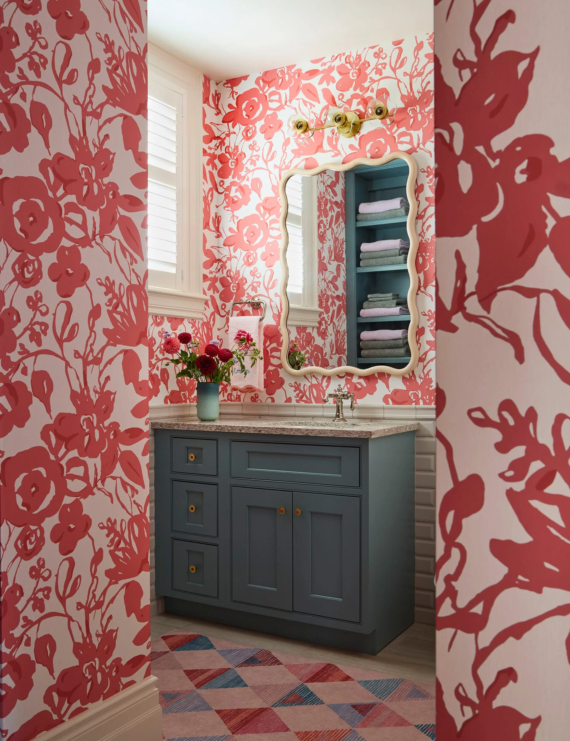 A powder room featuring bold red and white floral wallpaper, a blue-gray vanity cabinet with a stone countertop, a wavy-edged mirror, and recessed shelves.