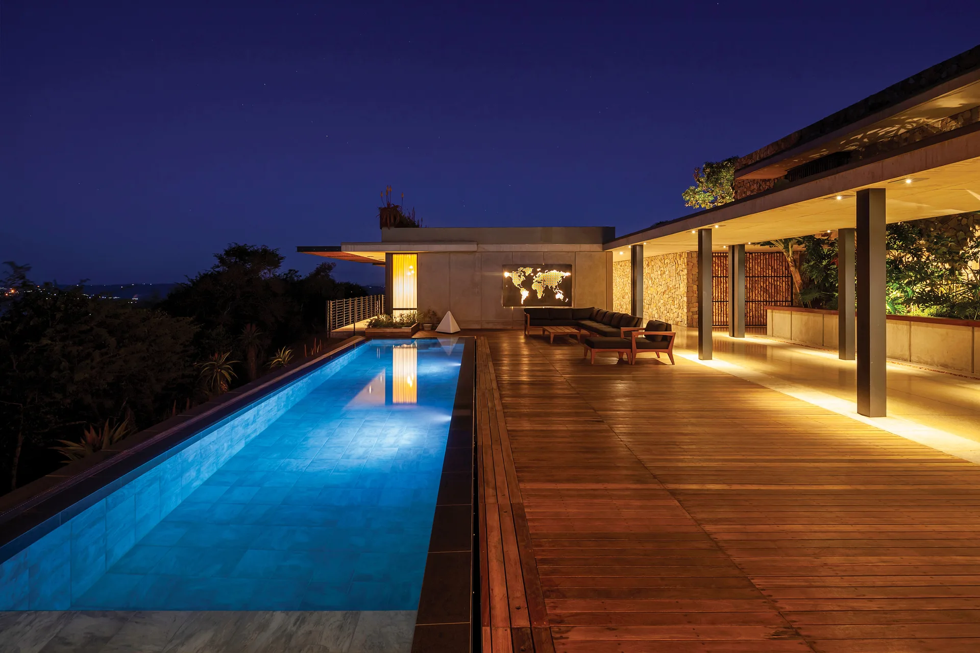 A long lap pool illuminated with blue light next to a spacious wood plank deck and a covered modern patio with recessed lighting.