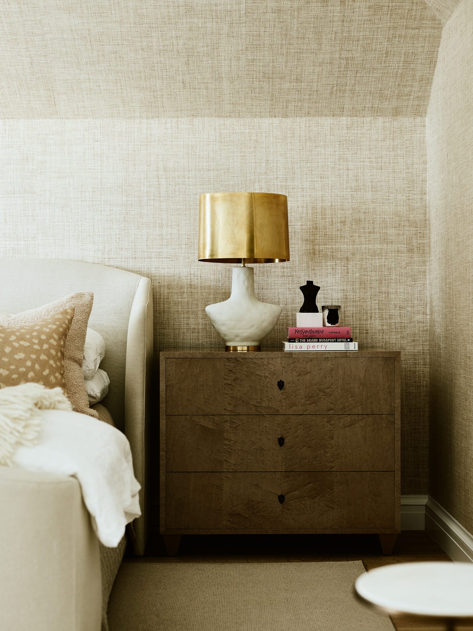 A bedroom corner features a wooden nightstand with a brass lamp in front of a textured beige grasscloth wallpapered wall next to an upholstered bed.
