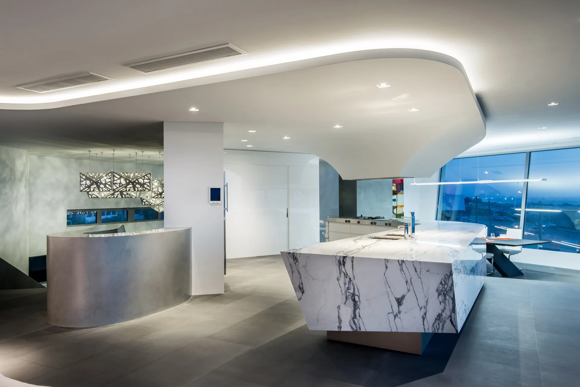 A modern kitchen features a large veined marble island, a curved metal bar, and a sculptural dropped ceiling with integrated cove lighting.