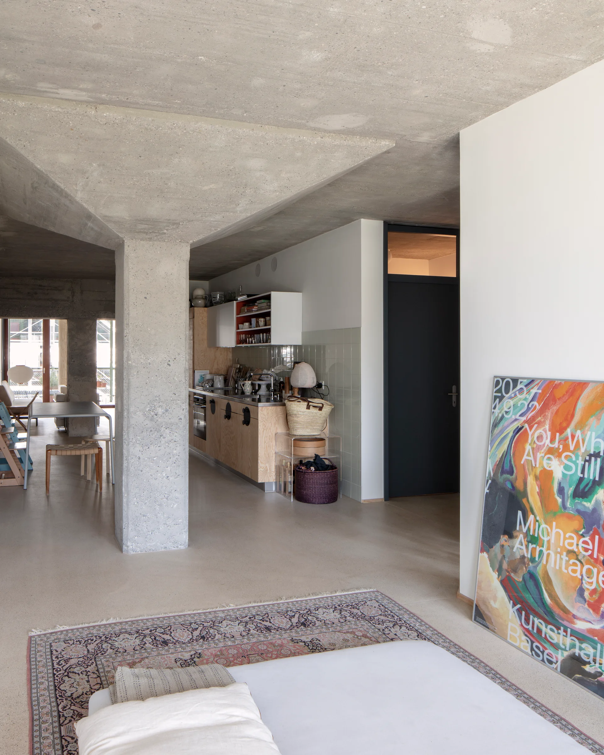An open-plan kitchen featuring plywood cabinets, a raw concrete column and ceiling, a traditional patterned rug, and a large art poster leaning on a white wall.