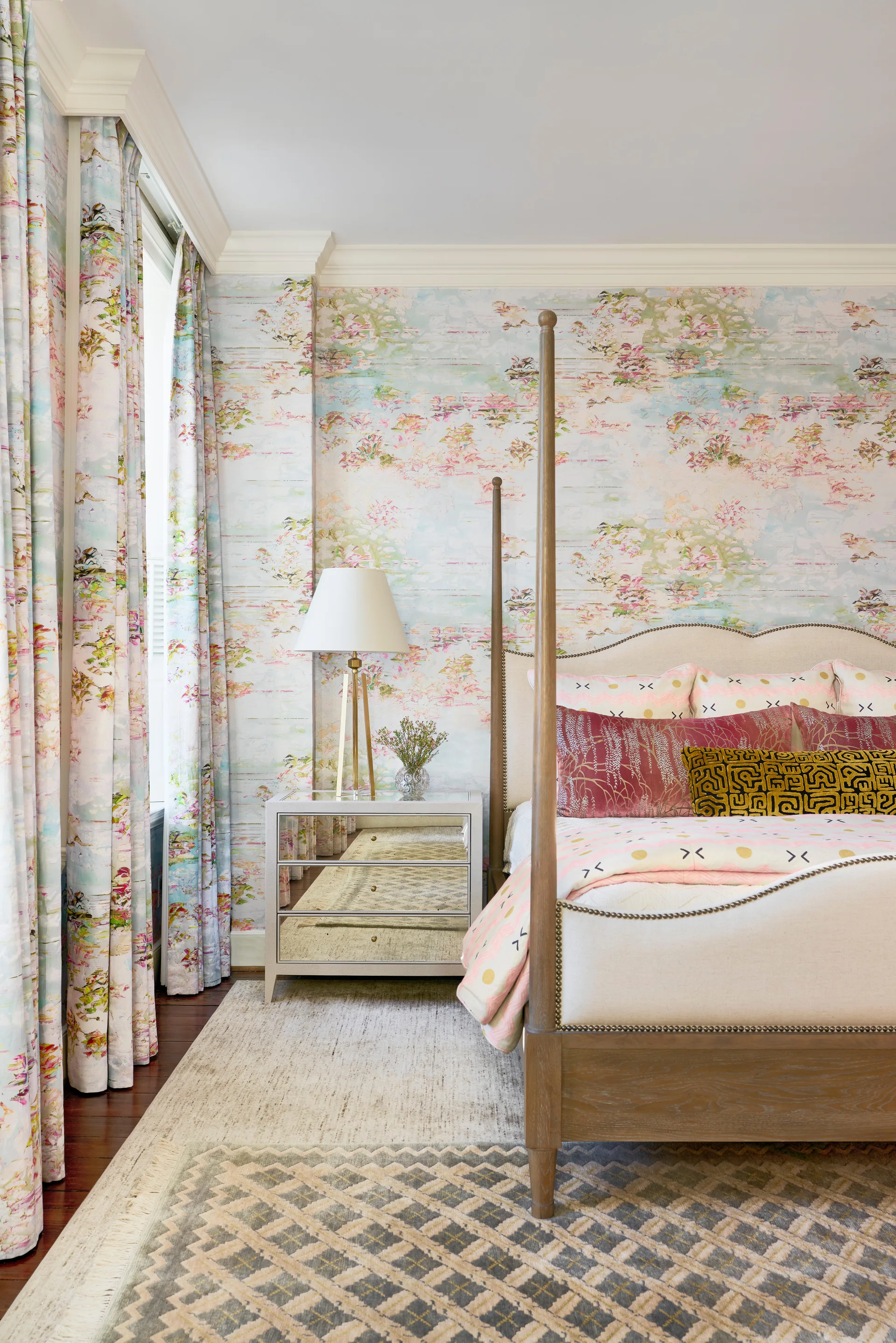 A bedroom featuring a wooden four-poster bed, a mirrored three-drawer nightstand with a lamp, and matching pastel scenic wallpaper and curtains.