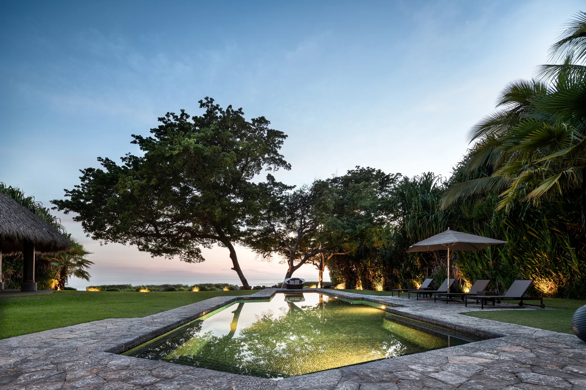 A rectangular swimming pool with irregular stone decking is surrounded by a green lawn, mature trees, and poolside lounge chairs at sunset.