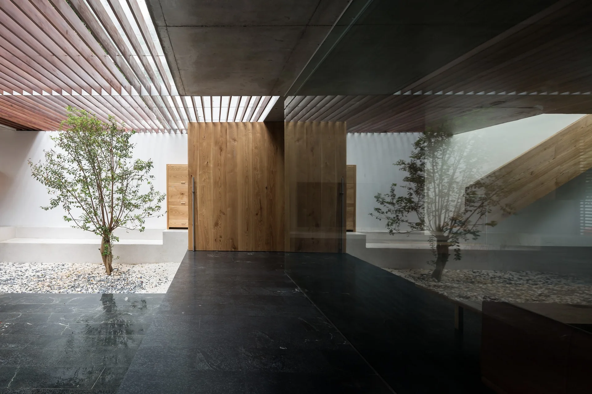 An architectural entryway features a slatted wood ceiling, an indoor garden with a small tree, and polished dark stone tile flooring.