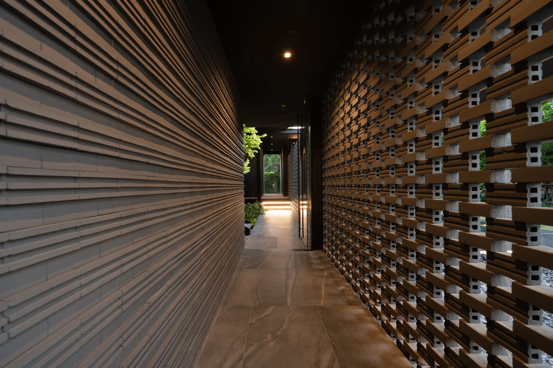 A dark modern hallway features a textured gray wall on the left and a breeze block masonry wall on the right with stone tile flooring.