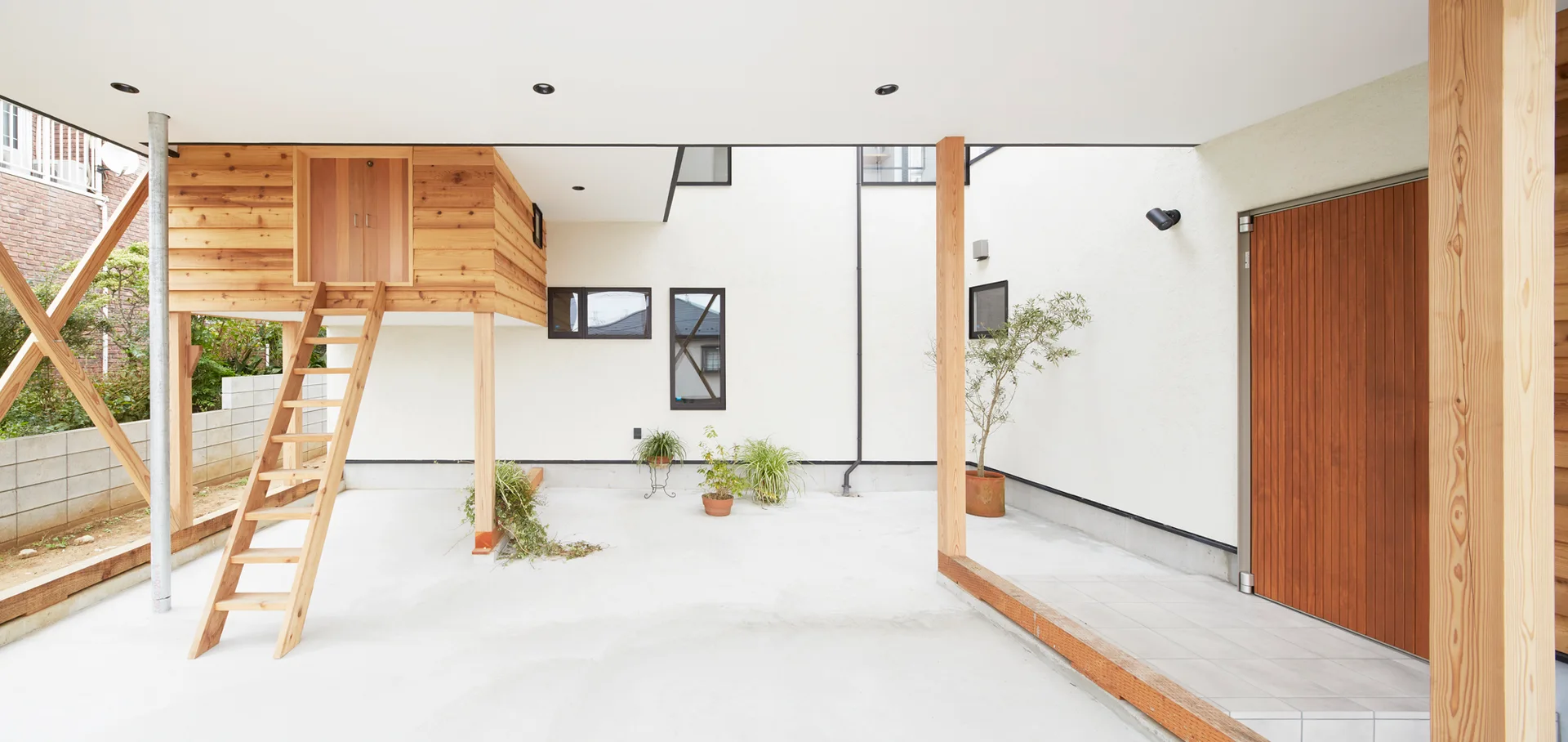 A covered outdoor patio with concrete flooring, a minimalist white wall, a tall wooden door, and an elevated wooden structure with a ladder.