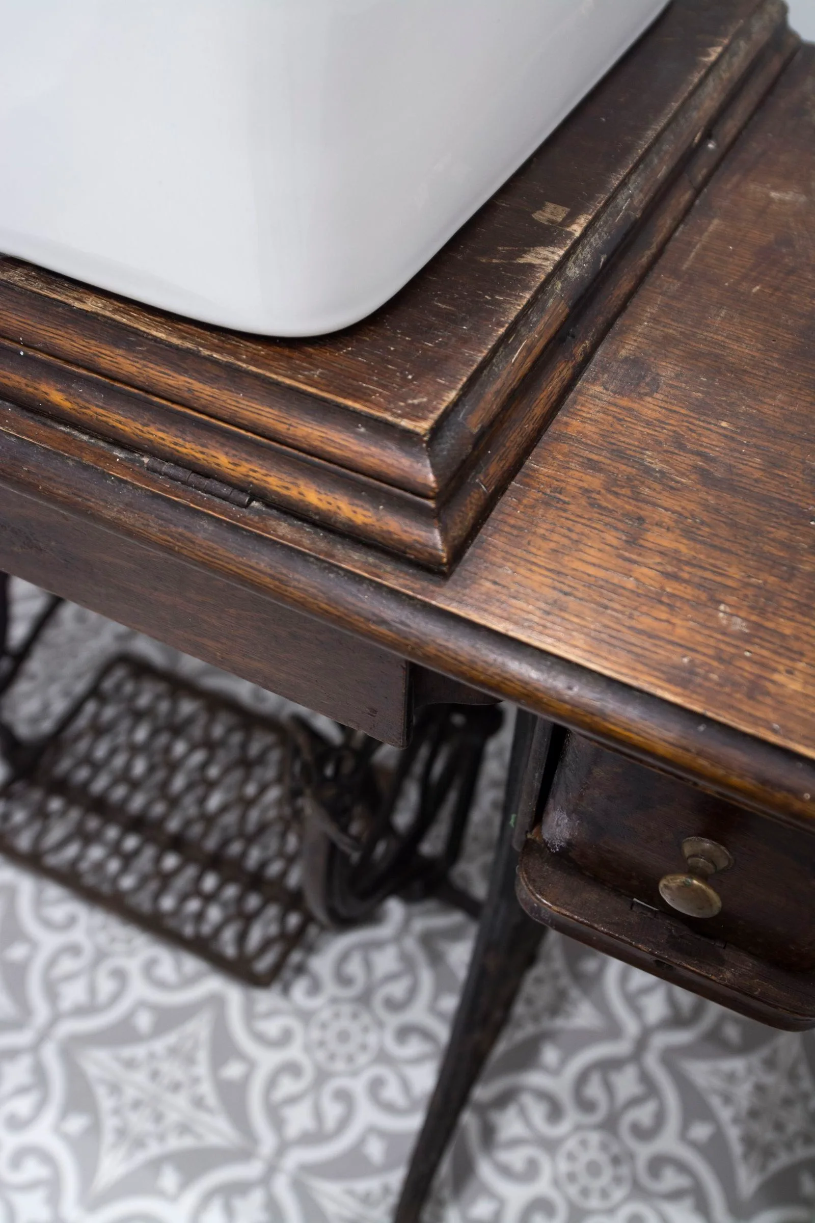 A close-up of a white ceramic sink basin mounted on a dark wood antique sewing machine table over grey and white patterned floor tiles.