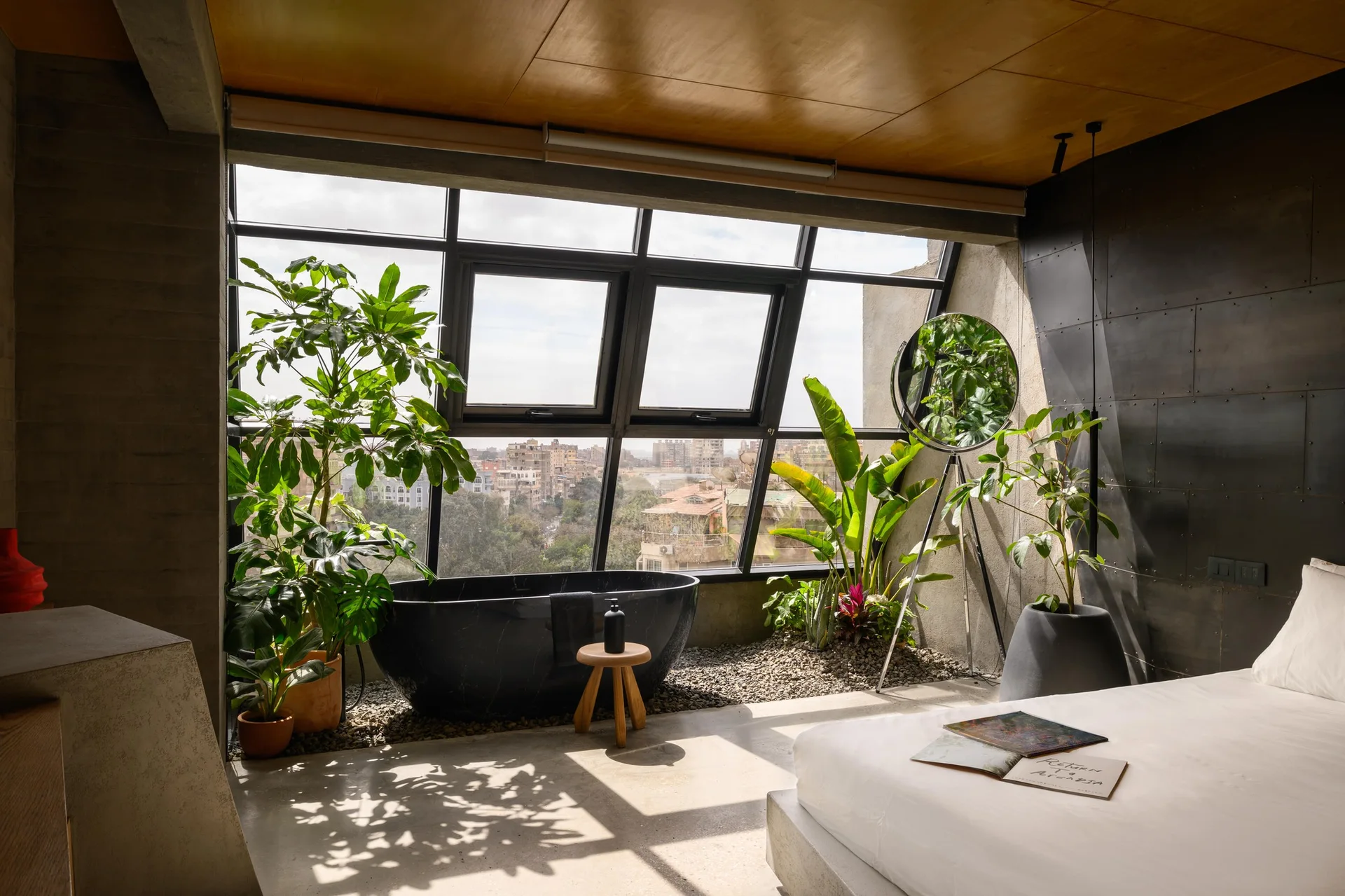 A modern industrial bedroom features a black freestanding bathtub on an indoor gravel bed next to a large window overlooking a city.