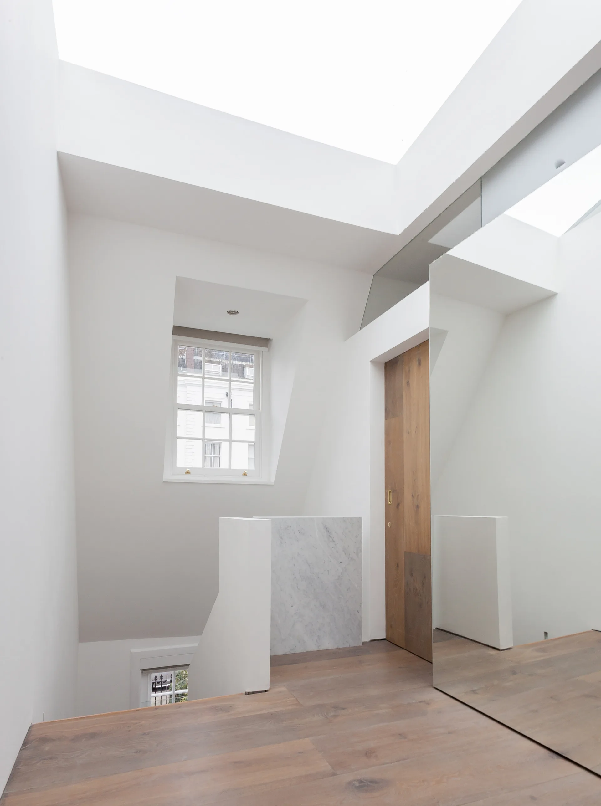 A bright minimalist stair landing features wood flooring, a tall wooden door, a recessed sash window, a marble accent wall, and a large skylight overhead.
