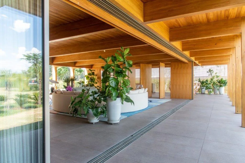 A covered outdoor patio featuring a warm timber ceiling with exposed beams, a curved white sofa, grey tile flooring, and tall potted plants.