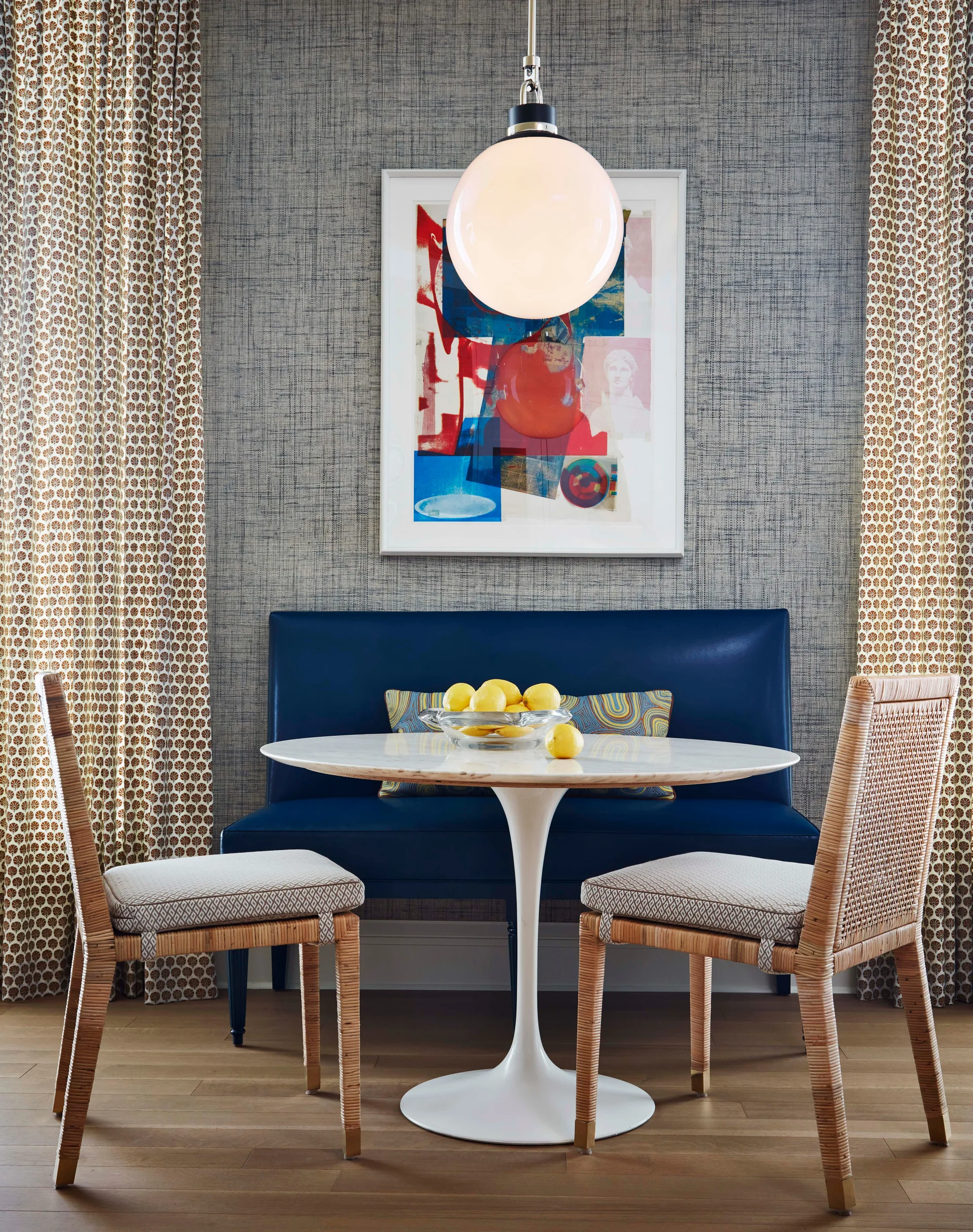 A dining nook with a blue leather bench, a white pedestal table, two rattan chairs, and patterned curtains against textured wallpaper.