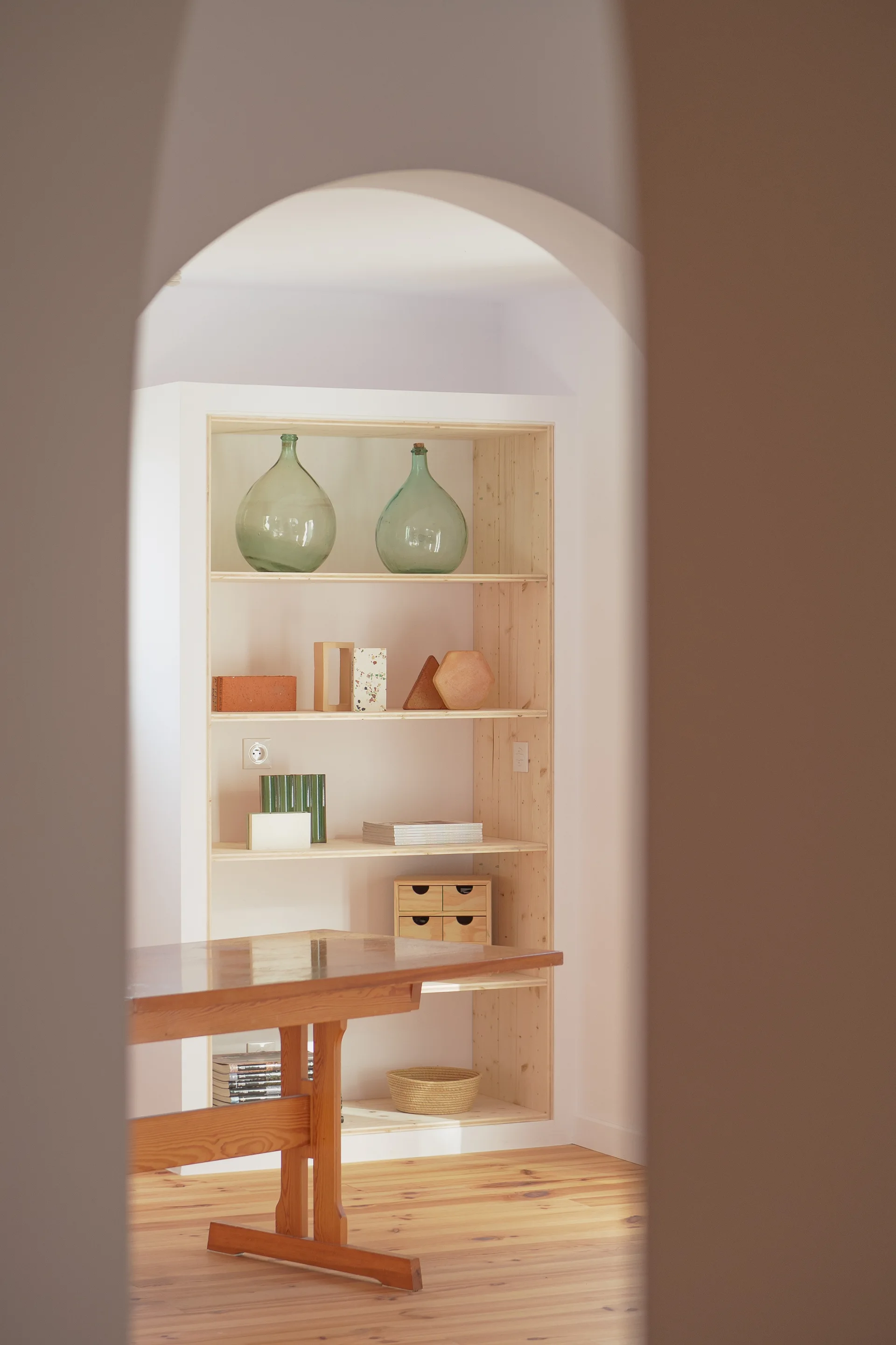An arched opening frames a light wood built-in shelving unit styled with green glass bottles and geometric ceramic objects next to a wooden table.