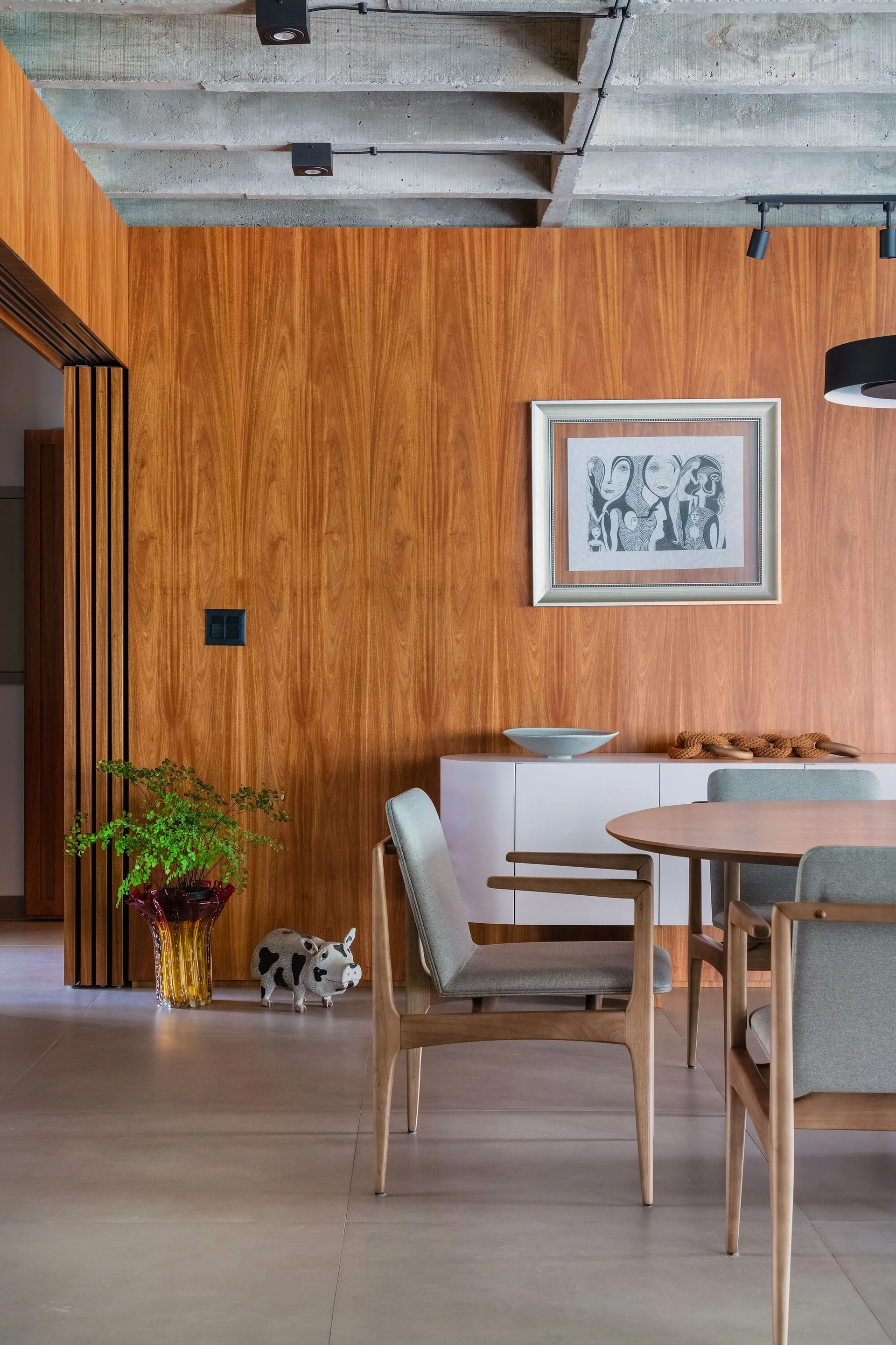 A modern dining room featuring a round wooden table and grey chairs against a vertical wood grain paneled wall beneath an exposed concrete waffle ceiling.