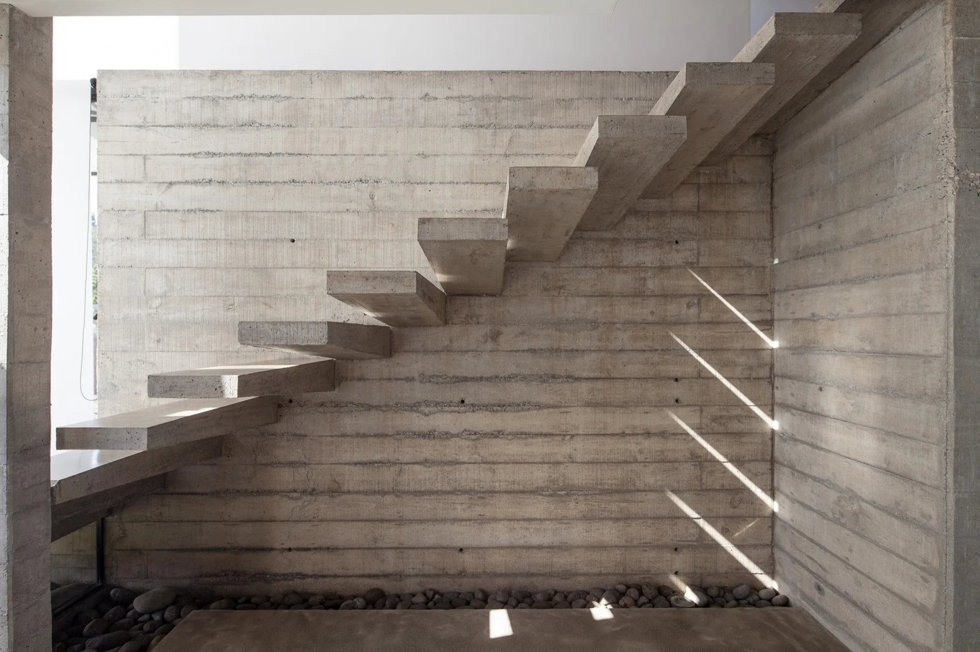 A minimalist cantilevered concrete staircase extends from a board-formed concrete wall with a bed of river stones at the base.