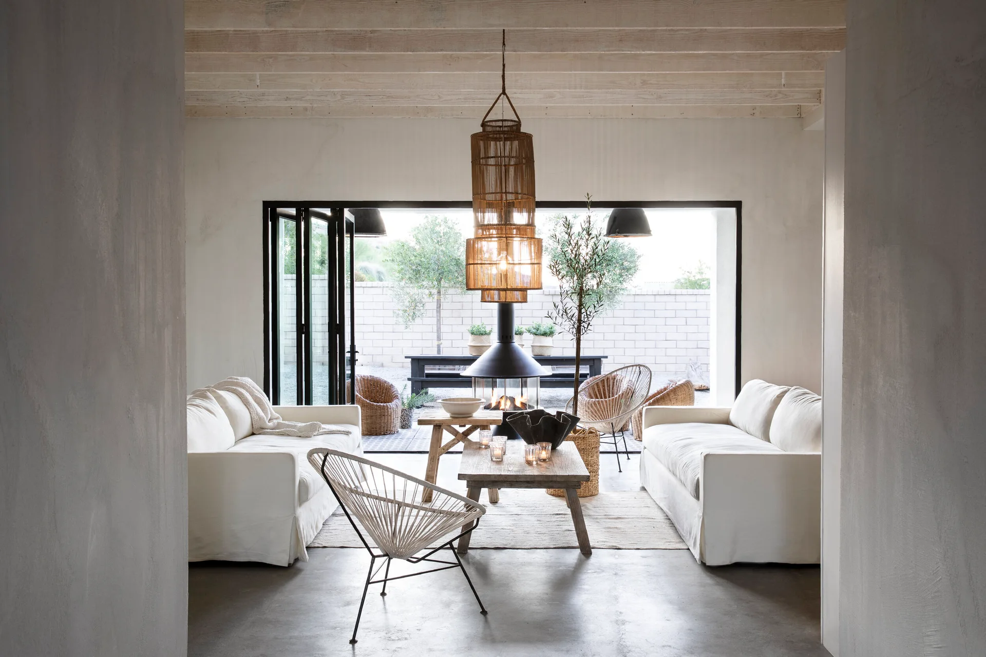 A light-filled living room featuring two white sofas facing a rustic wood coffee table, with a black suspended fireplace in front of open bi-fold doors.