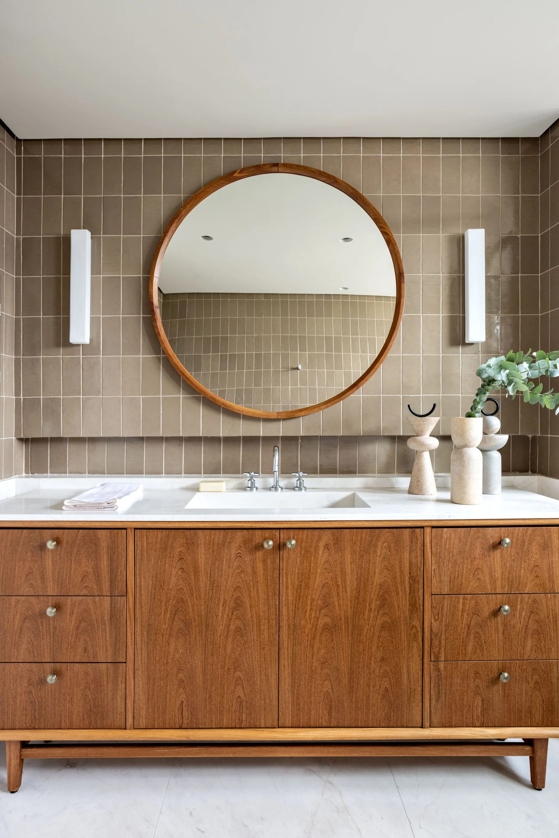 A bathroom vanity with a wood cabinet, white marble countertop, a large round wooden mirror, and vertical white sconces against a taupe tiled wall.