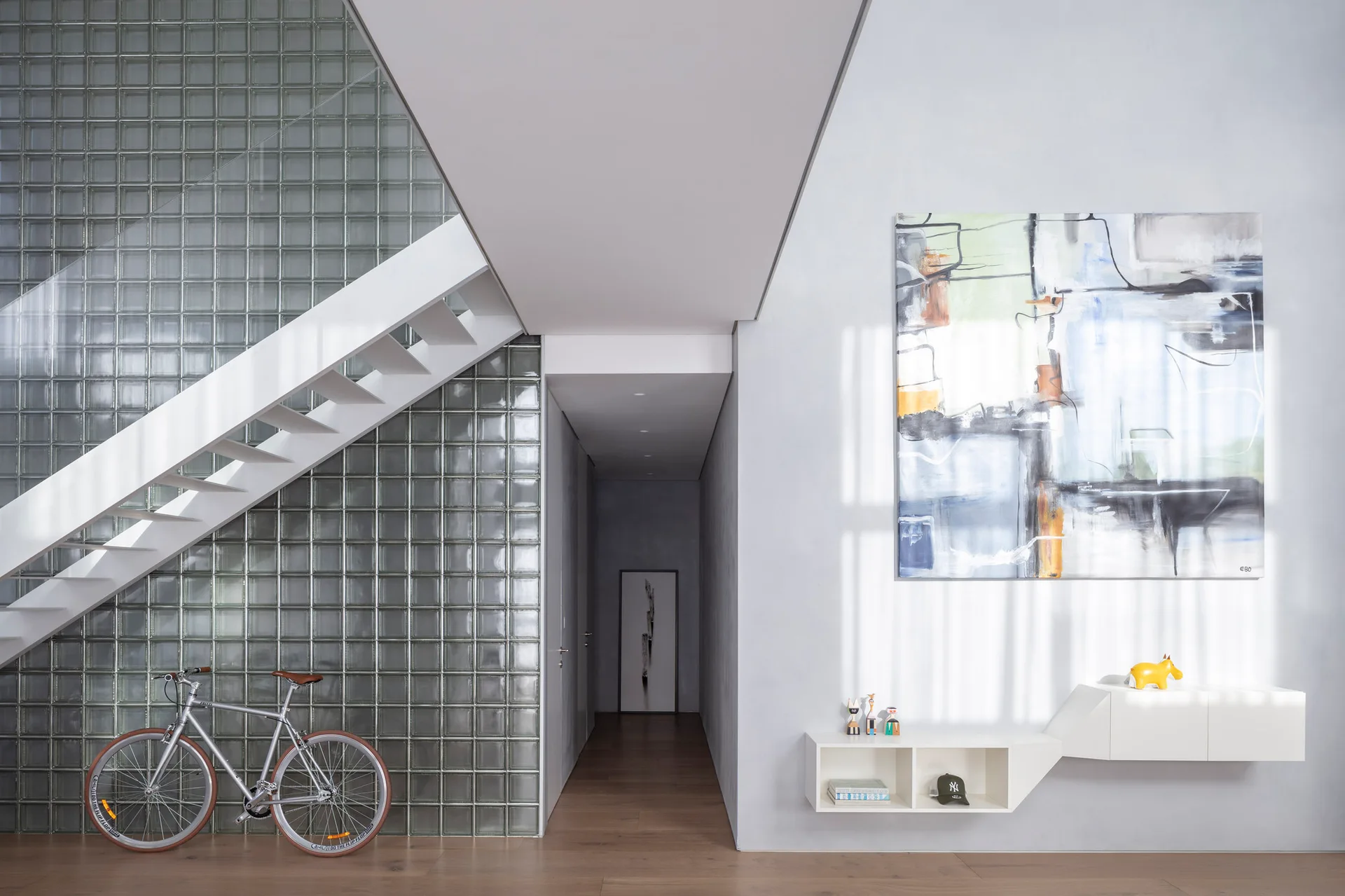 A white floating staircase climbs in front of a glass block wall next to a grey hallway with a wall-mounted console and abstract artwork.