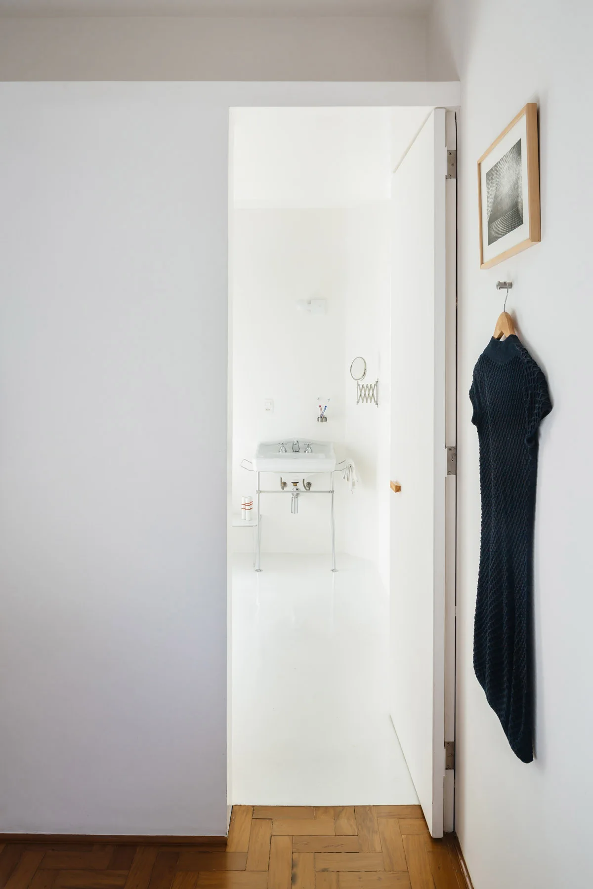 A view through an open doorway from a wood parquet hallway into a minimalist white bathroom with a metal-legged console sink.