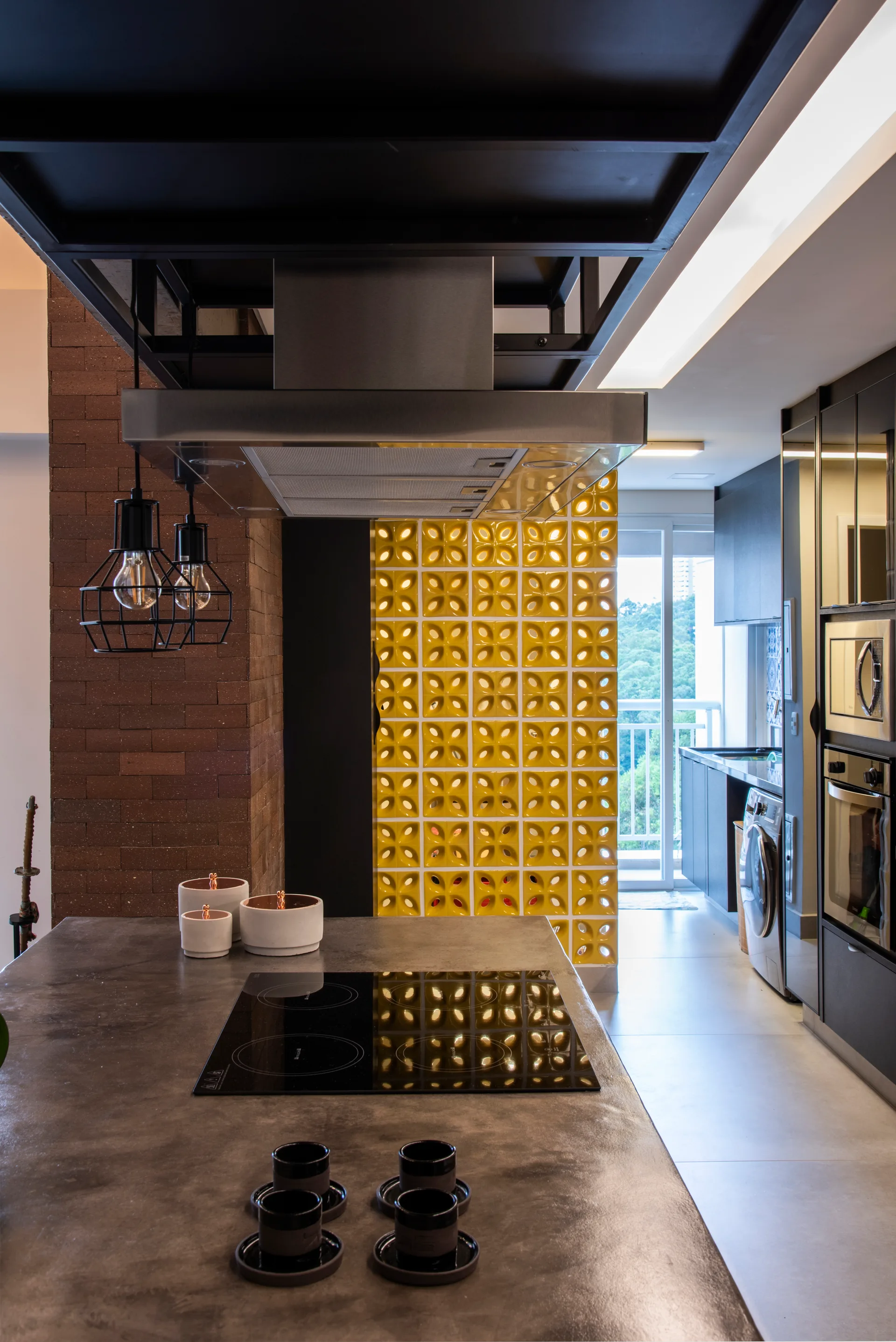 A modern kitchen featuring a concrete island with a cooktop, a dark brick pillar, and a bright yellow patterned breeze block partition wall.