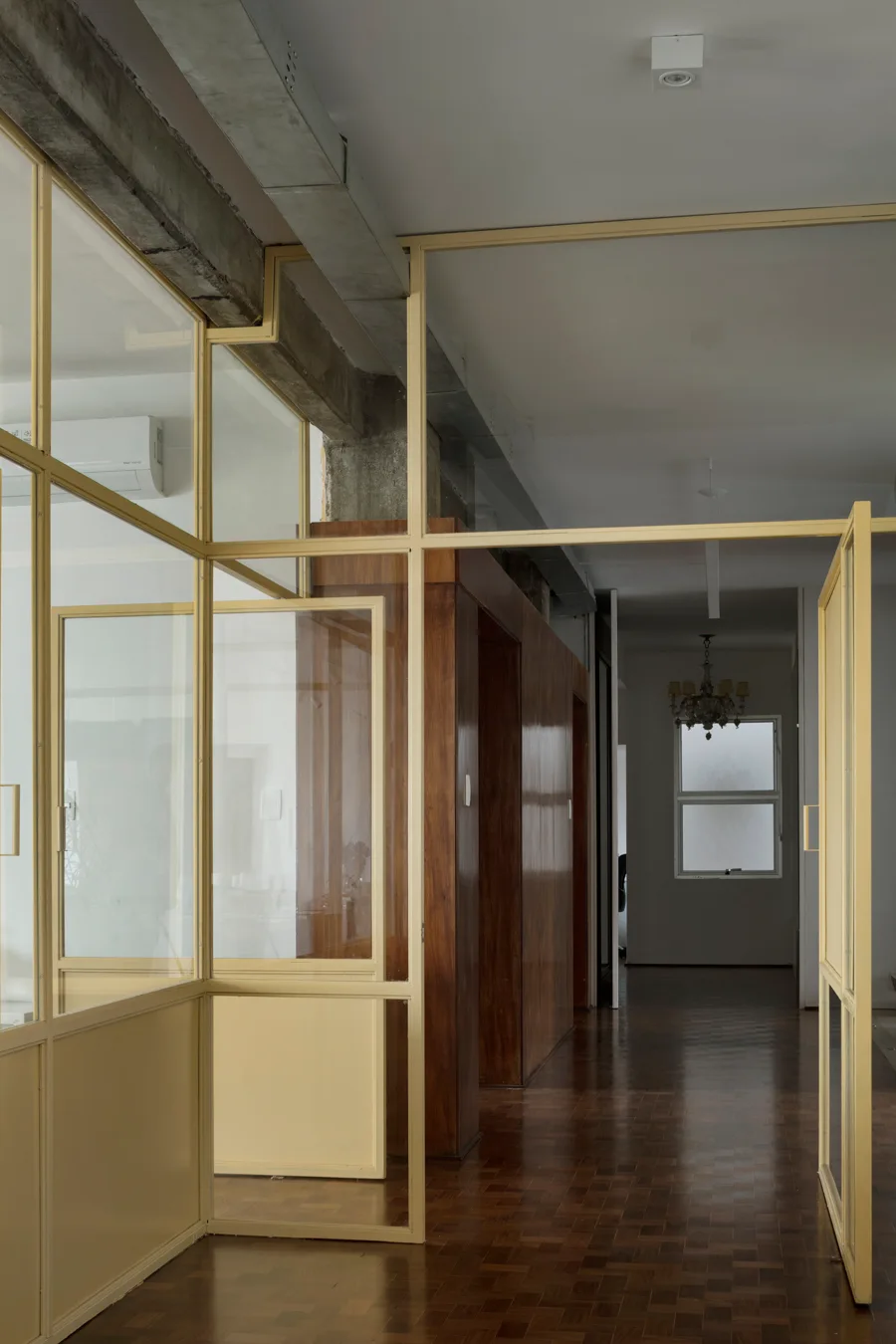 An interior hallway features a yellow-framed glass partition wall, warm wood parquet flooring, and an exposed concrete ceiling beam.
