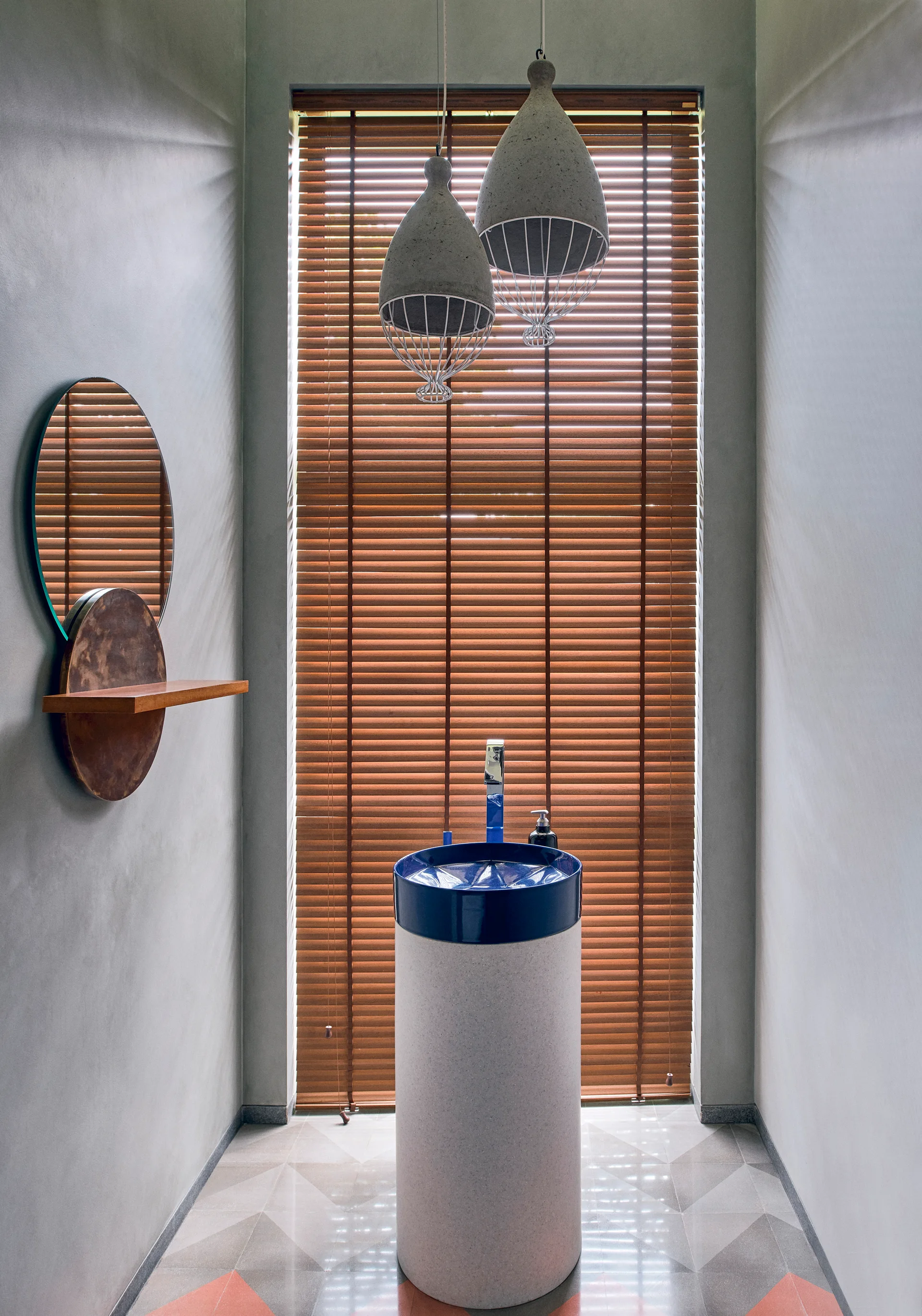 A modern powder room features a cylindrical pedestal sink with a blue basin, set against a tall window with brown wooden blinds.