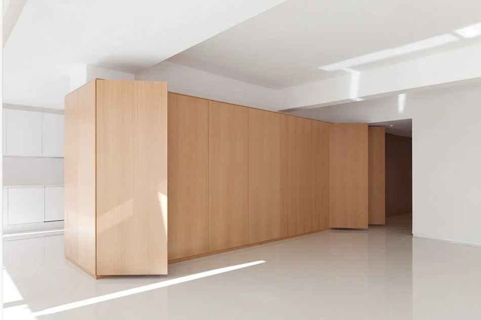A large floor-to-ceiling light wood cabinetry unit stands against white walls in a minimalist interior with reflective white flooring.