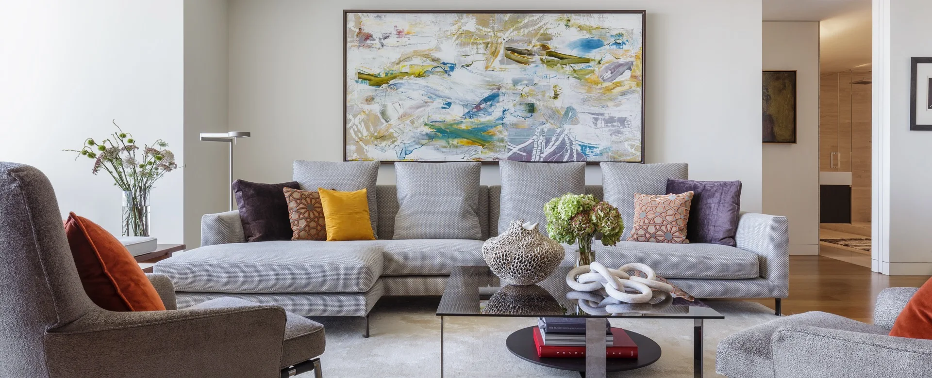 A grey textured sectional sofa features colorful accent pillows beneath a large abstract painting in a modern living room with a glass coffee table.