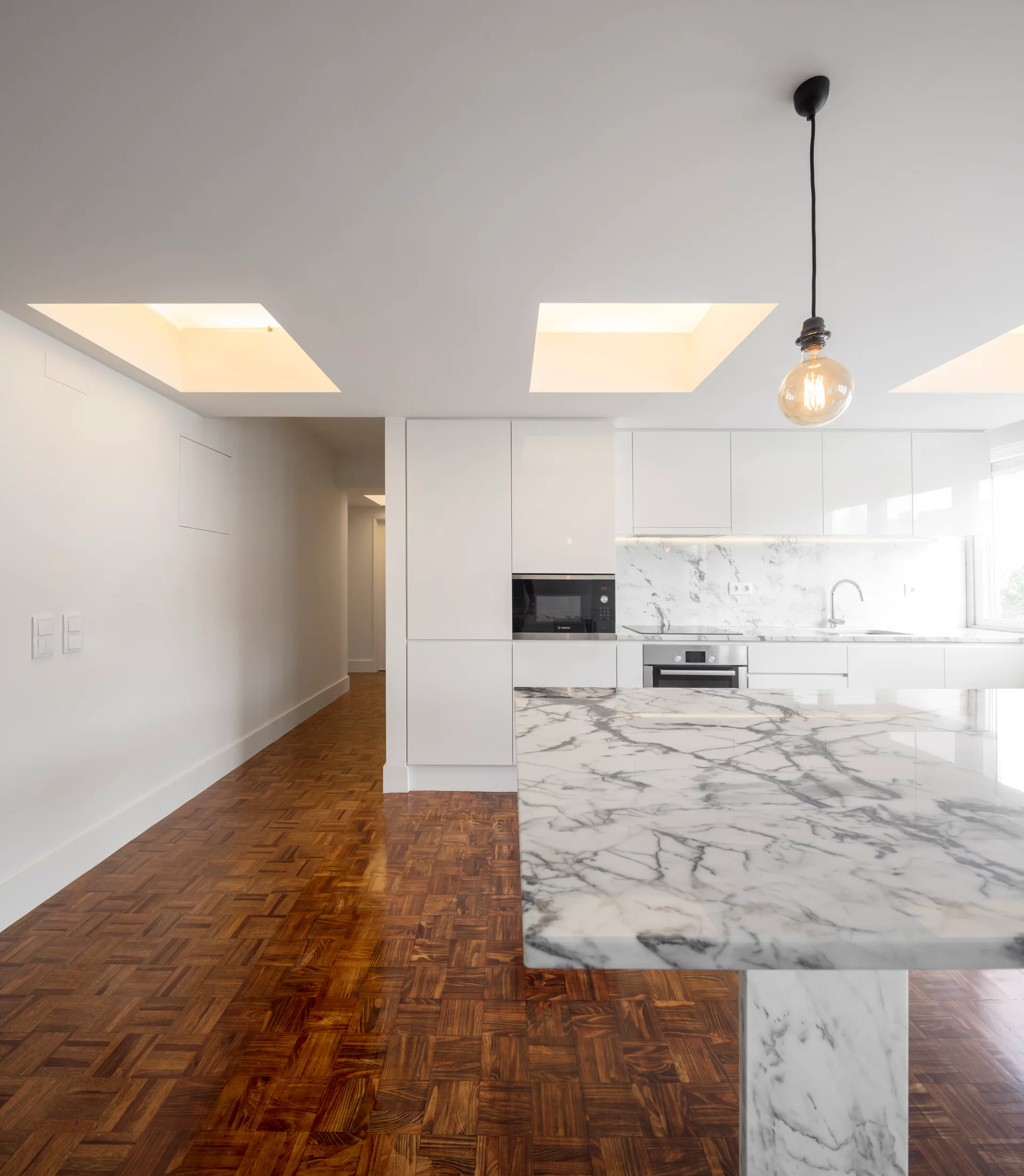 A minimalist kitchen features glossy white cabinetry, a large marble island with gray veining, and warm parquet wood flooring.