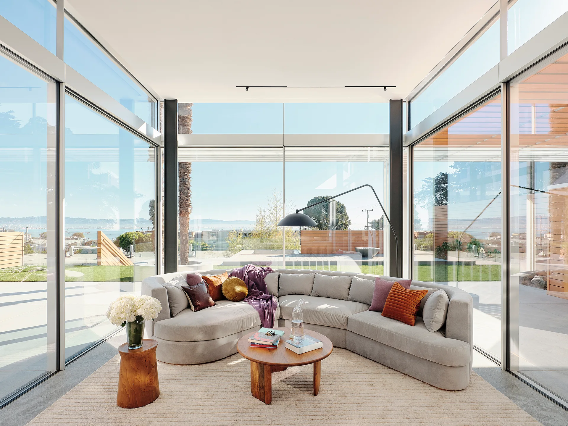 A light grey curved sectional sofa on a ribbed rug sits surrounded by floor-to-ceiling glass walls looking out to a patio.