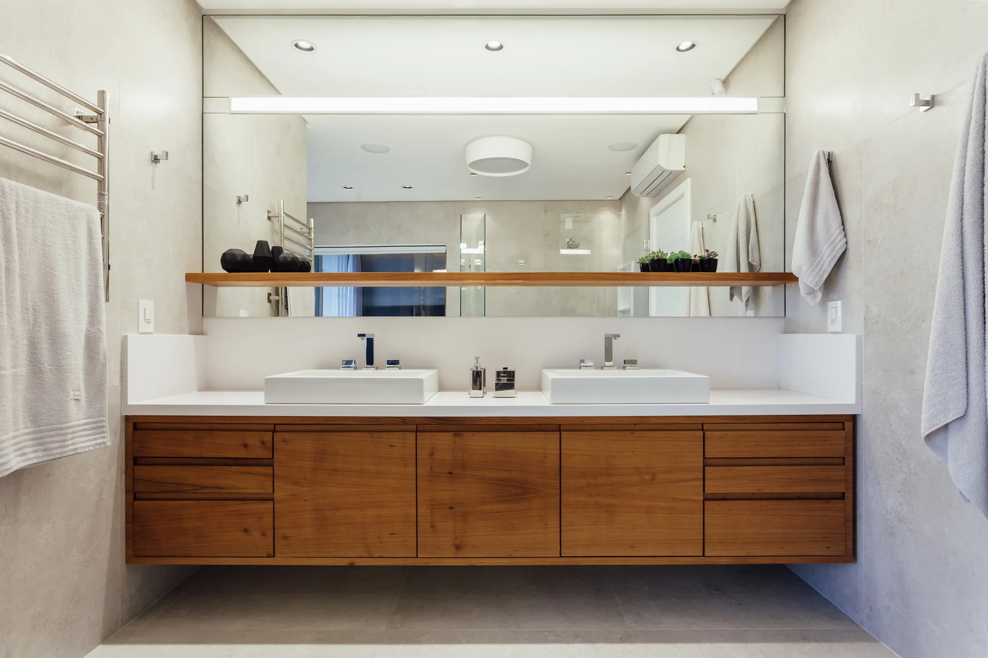 A modern bathroom with a floating natural wood vanity, two white sinks, a large mirror split by a wood shelf, and light grey tile walls.