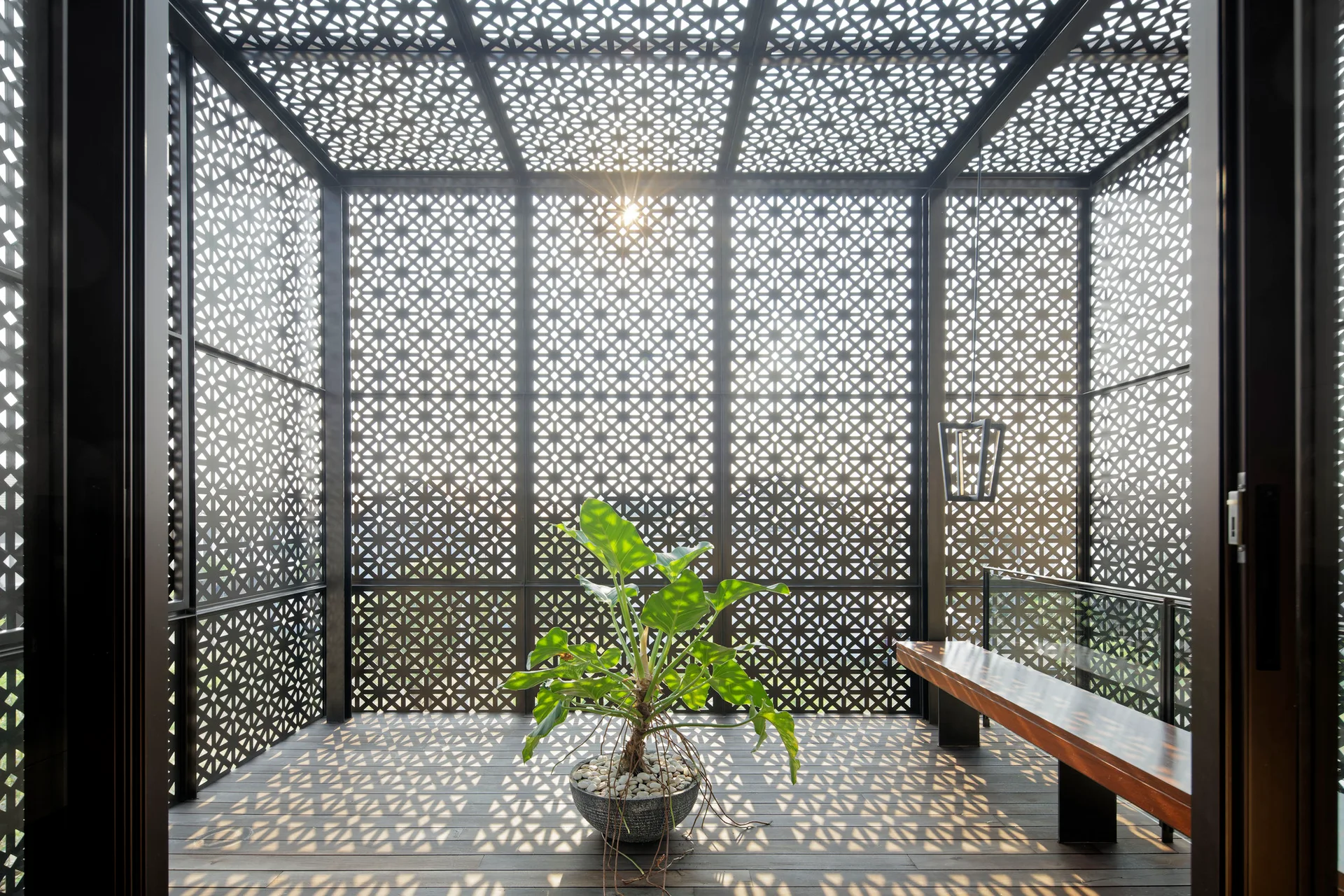 An enclosed outdoor terrace with black metal geometric lattice walls and ceiling, a single potted plant on the wood deck, and a wooden bench.
