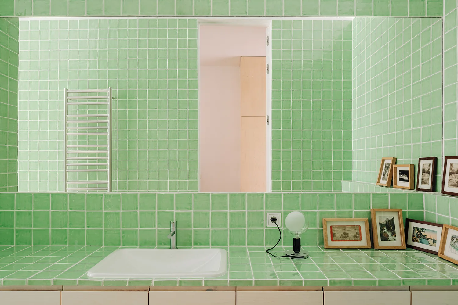 A bathroom vanity and wall covered in glossy green square tiles, featuring an integrated white sink, a large mirror, and small wooden picture frames.