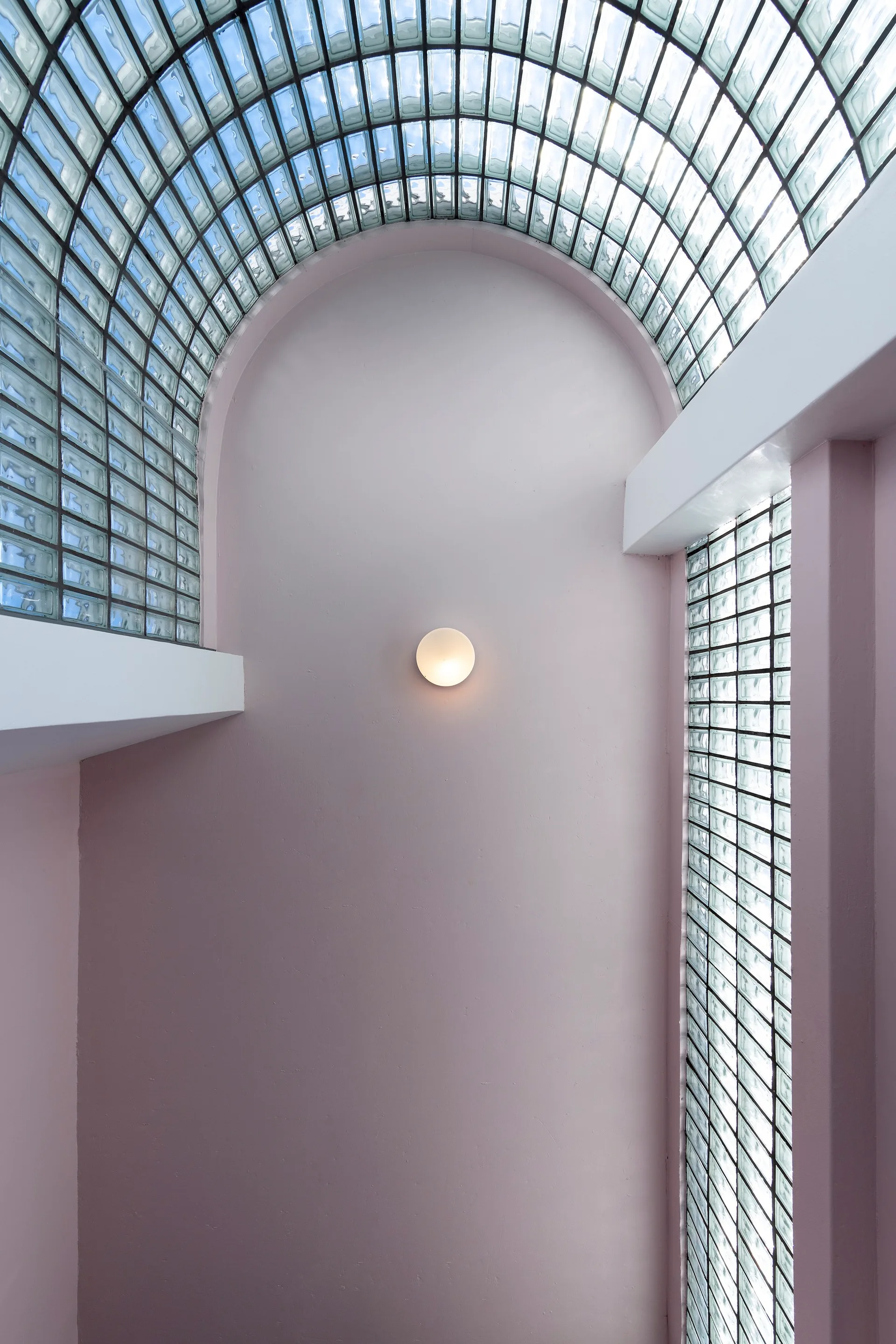 A curved blush pink wall is flanked by vertical glass blocks and topped with an arched glass block ceiling illuminated by a round sconce.