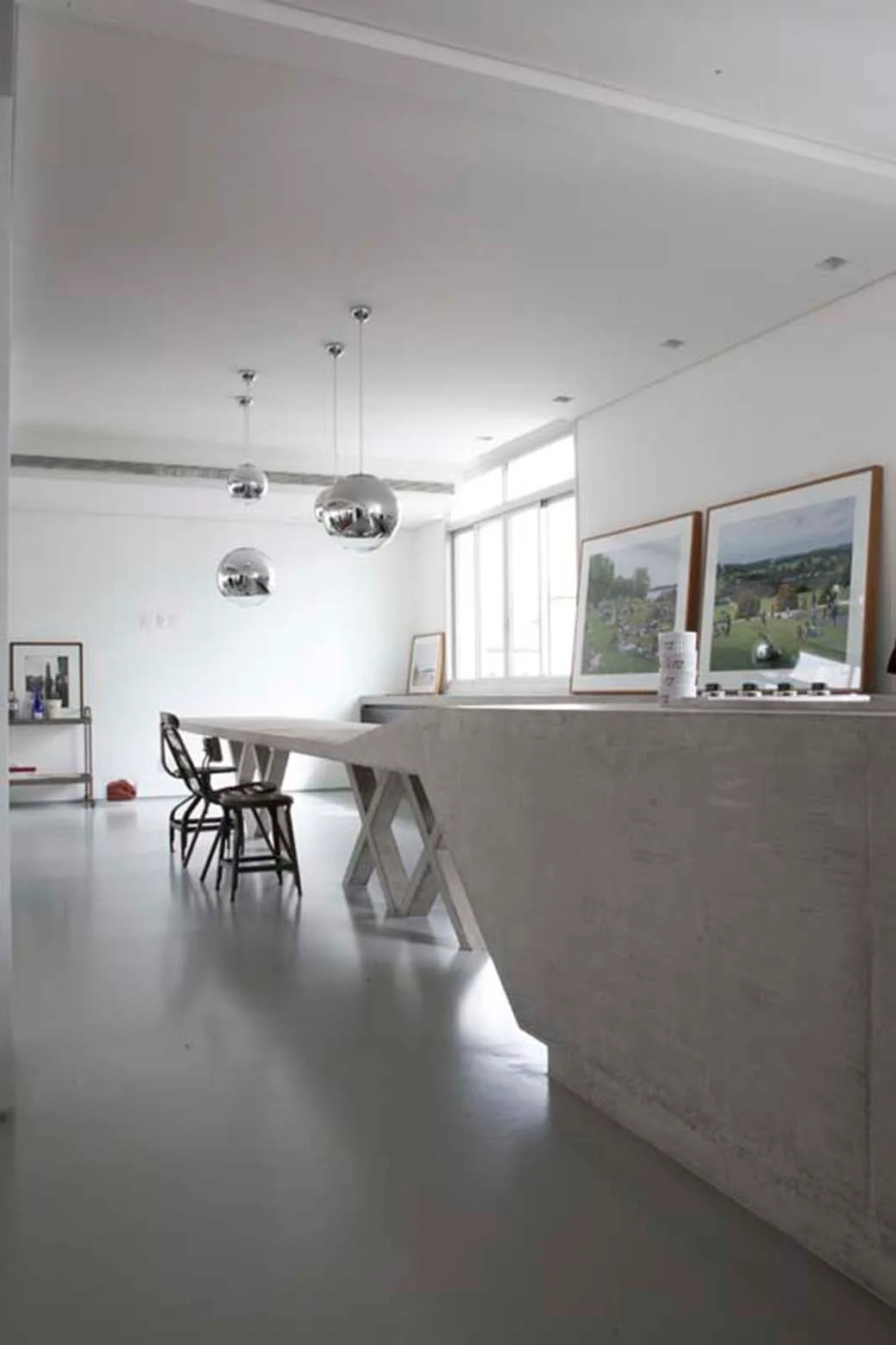 A minimalist industrial kitchen features a massive grey concrete island, black metal chairs, and spherical chrome pendant lights.