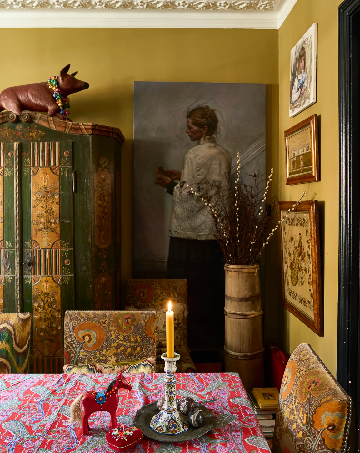 An eclectic dining room features a red paisley tablecloth, patterned upholstered chairs, olive green walls, and a tall painted wooden armoire.