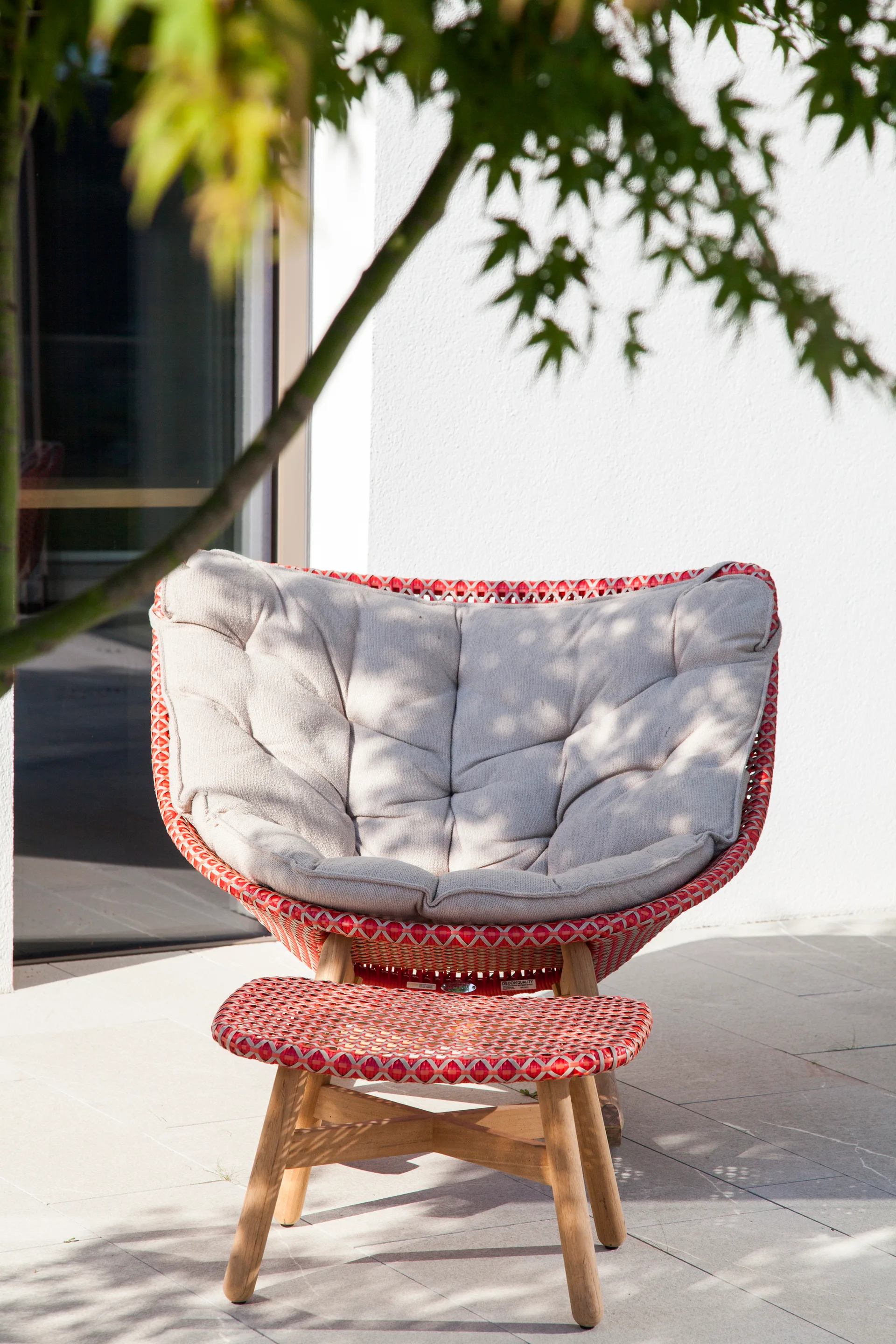 A red woven outdoor lounge chair with a beige cushion and a matching footstool sits on a paved patio under the shade of a tree.